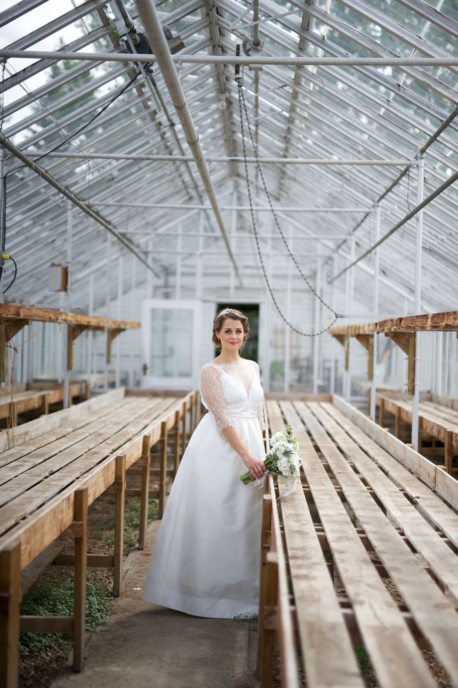 Bride looks over her shoulder in a rustic greenhouse, soft smile and lace gown catching the light.