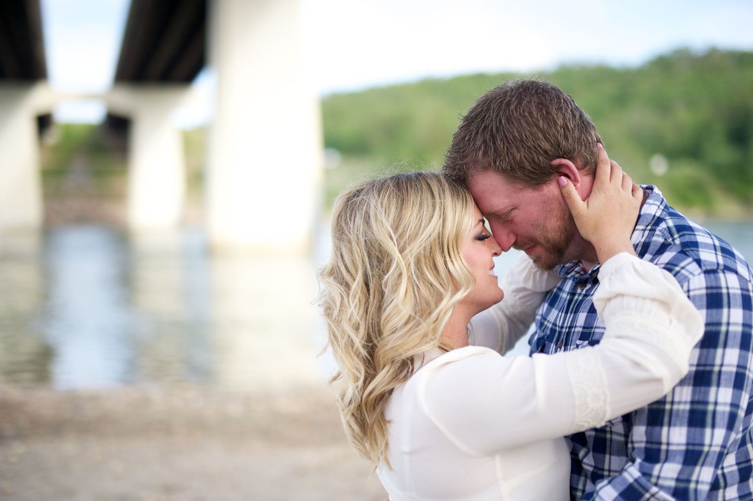 Engaged couple embracing near a riverbank, foreheads touching, captured under a bridge in golden light. Calgary engagement photo.