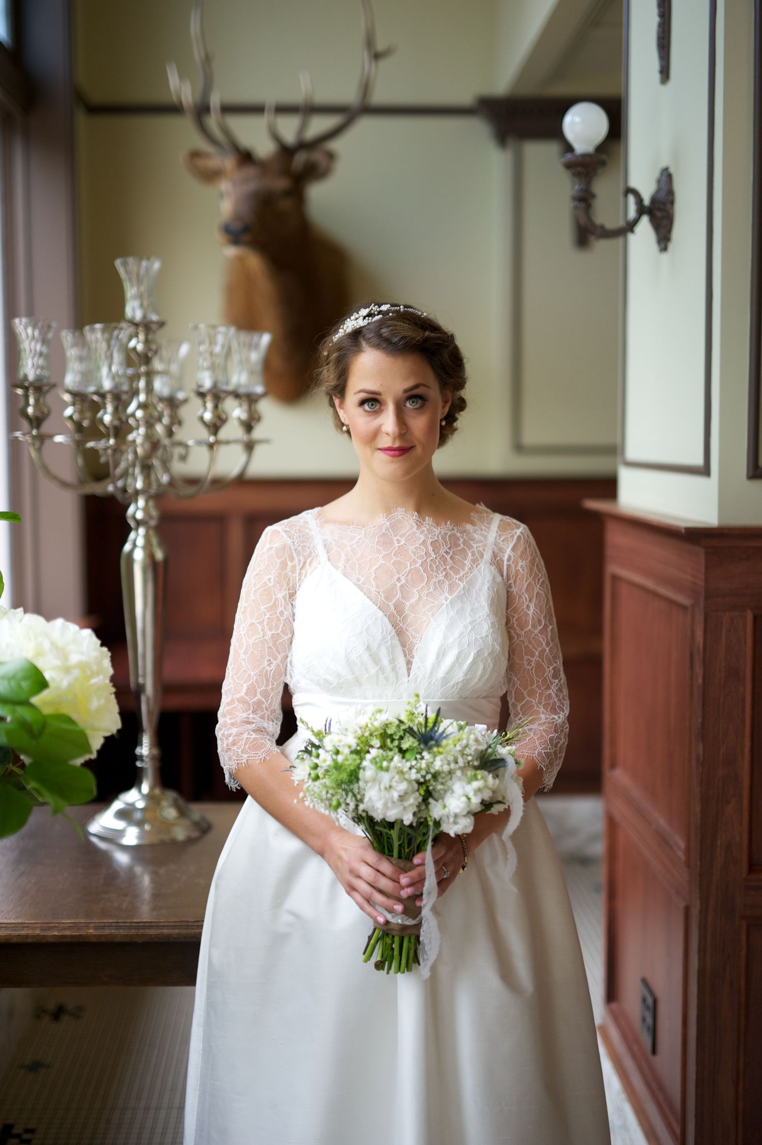 Bride stands softly by a window, bouquet in hand, framed by candlelight and antique decor.