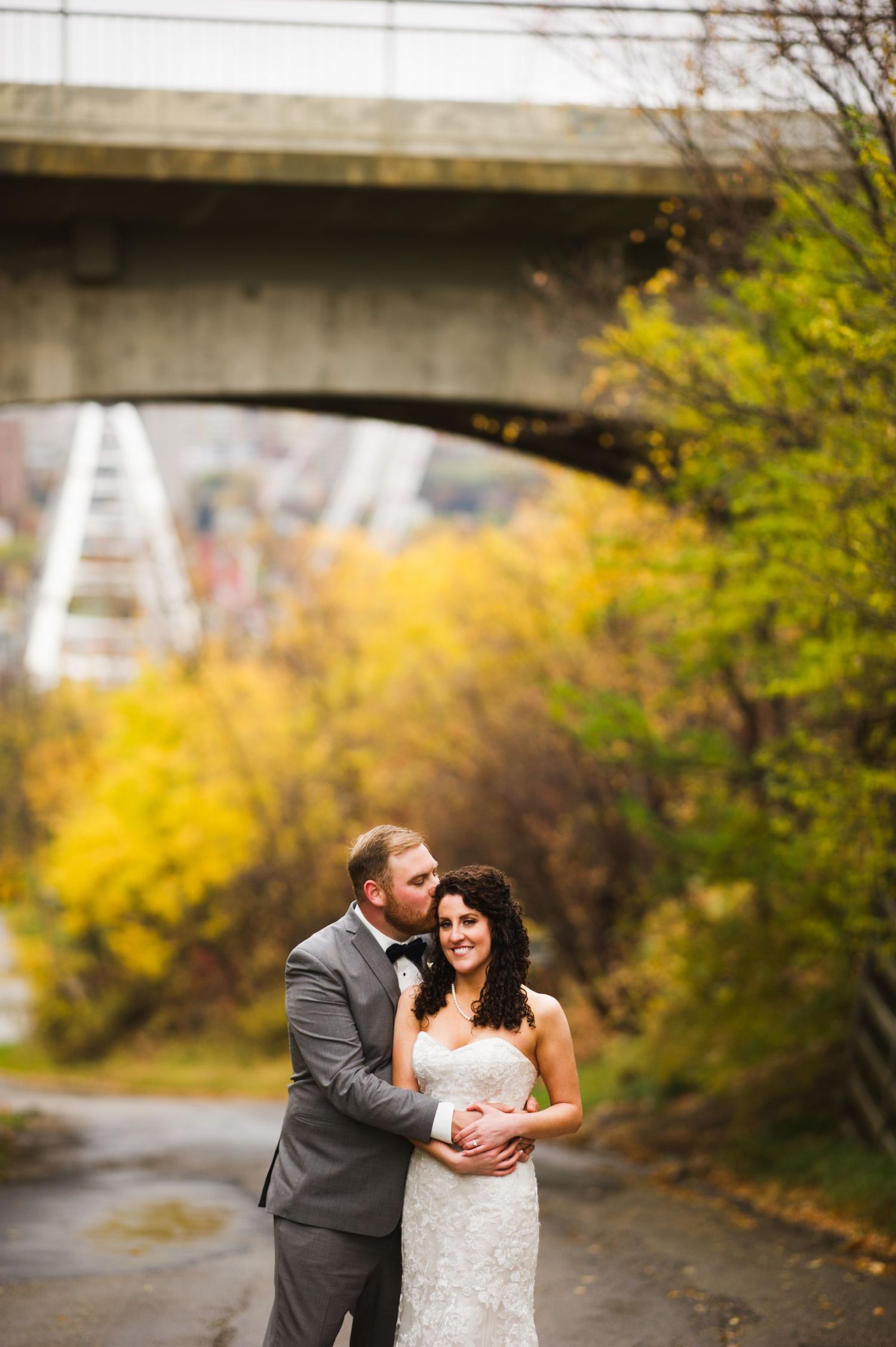 Groom gently kissing bride’s forehead as they stand together on a city pathway framed by colorful fall foliage and an overpass overhead.