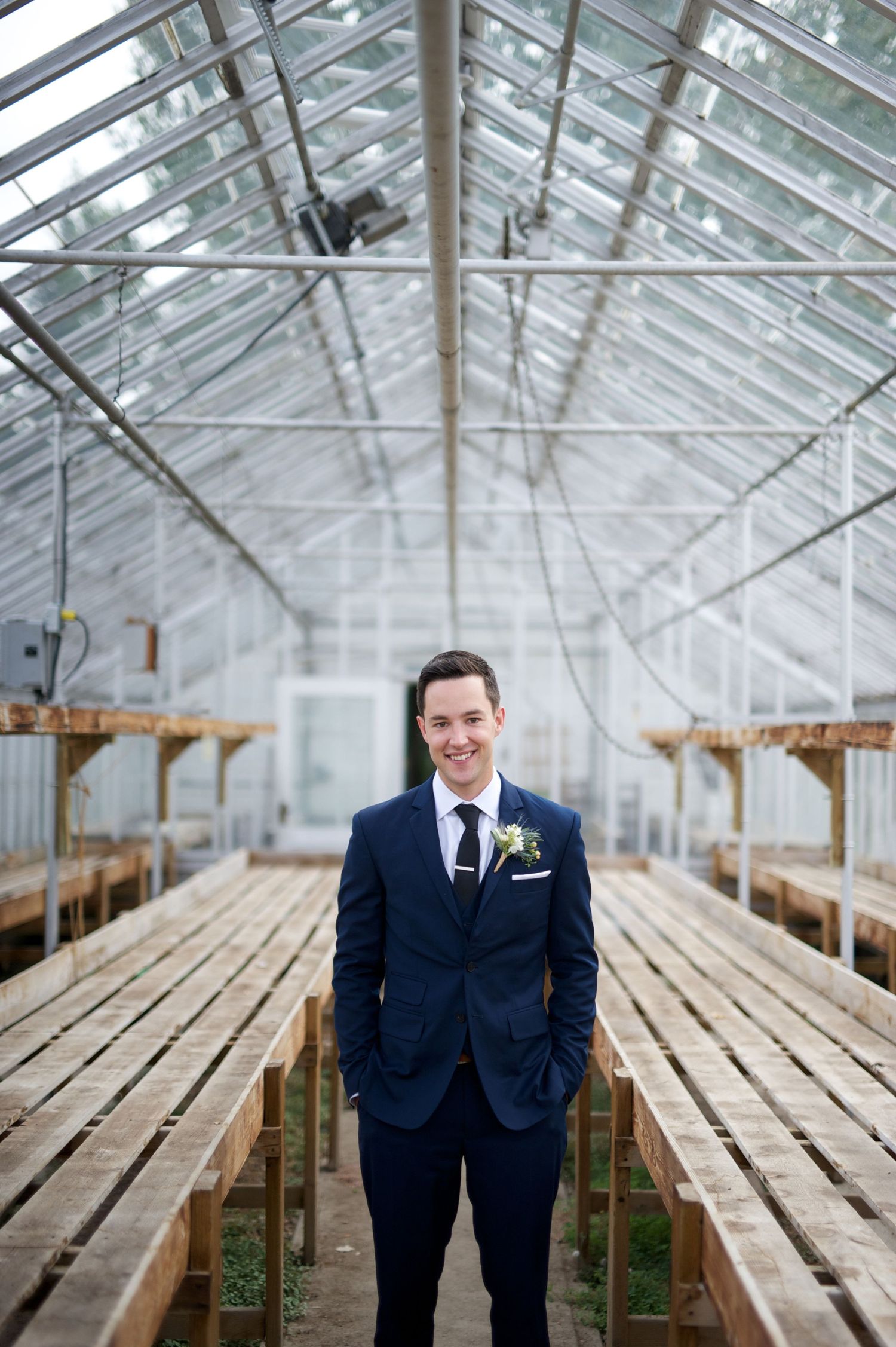 Groom stands in the center of a greenhouse, smiling with hands in pockets between long wooden planting tables.