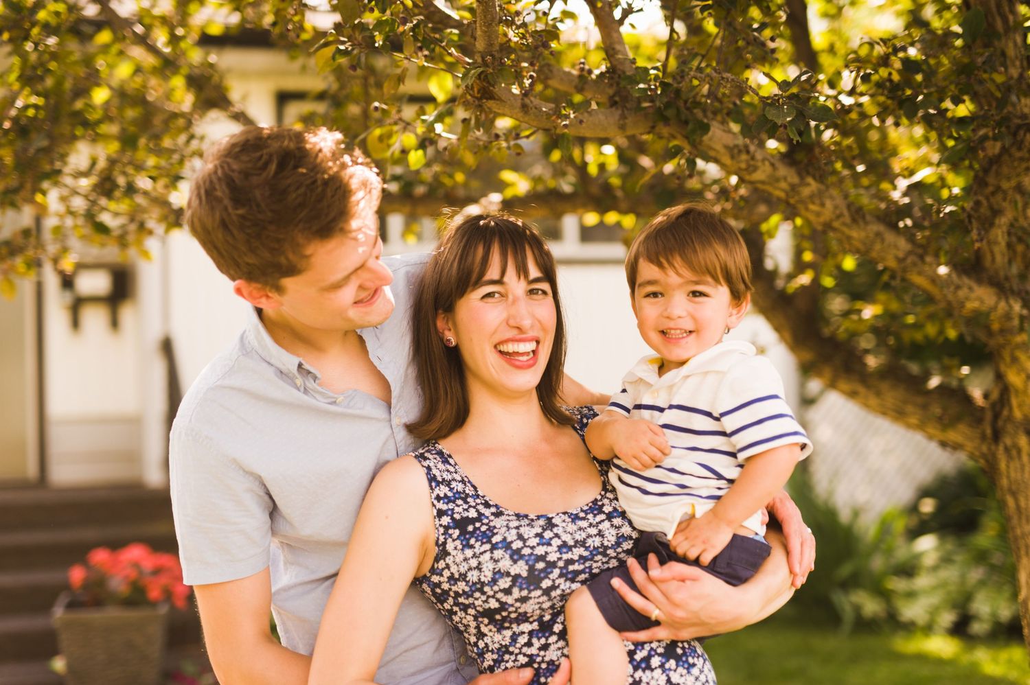 Parents and toddler laughing together under a backyard tree, sharing a joyful moment.