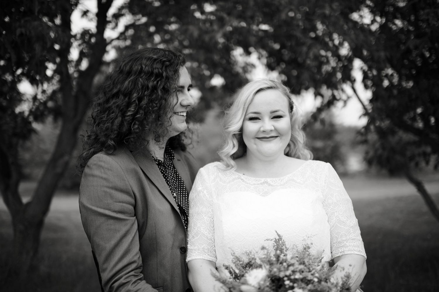 Black and white photo of newlyweds standing under trees, bride holding bouquet and smiling while groom gazes at her.