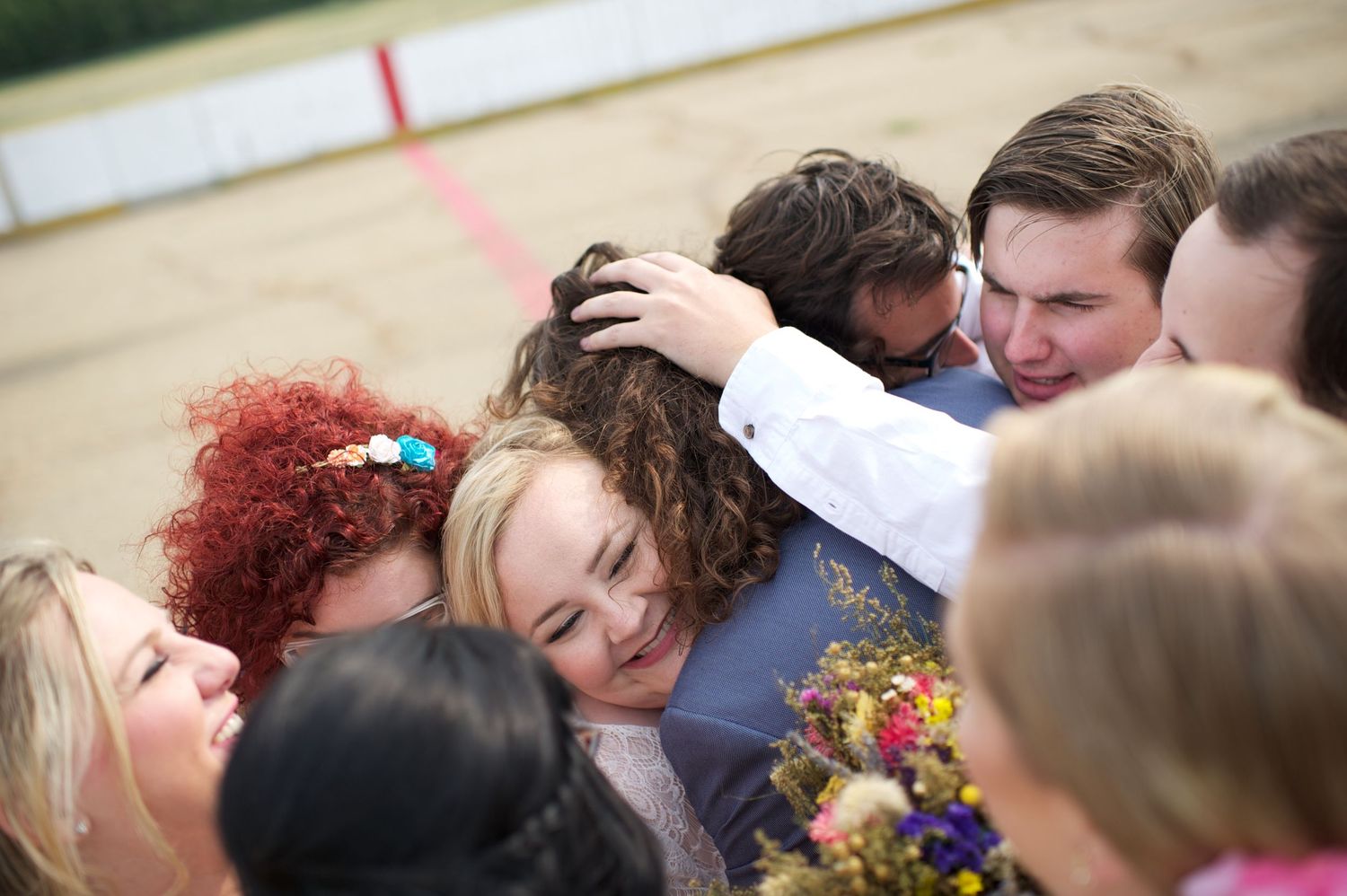 Bride and groom surrounded by their wedding party in a joyful group hug on an outdoor hockey rink. Genuine and emotional celebration moment.