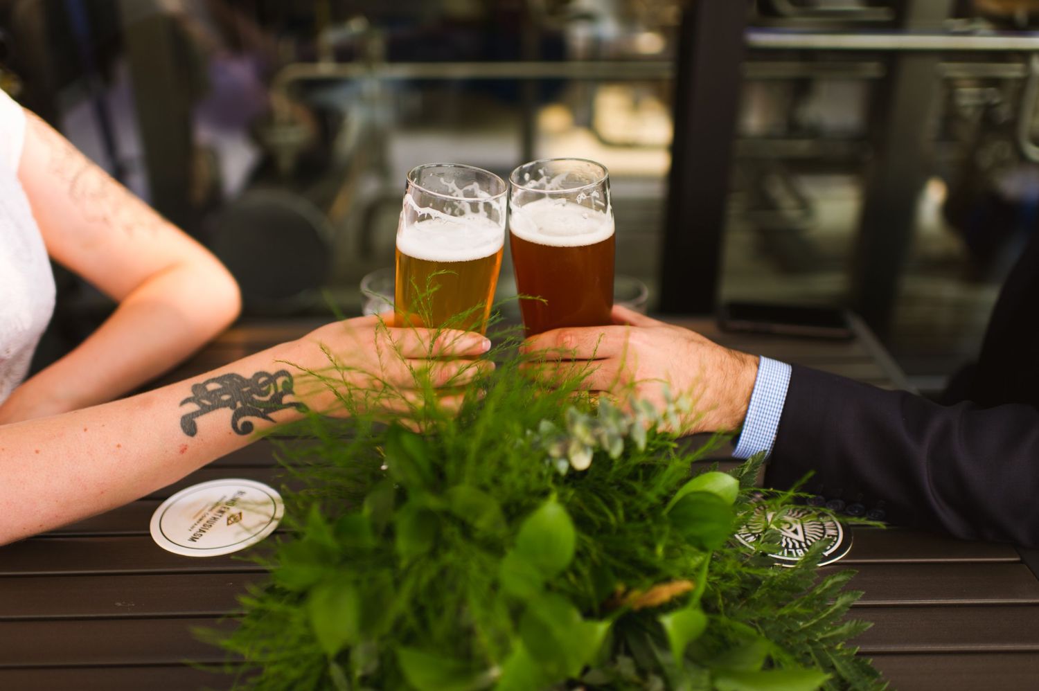 Close-up of couple’s hands and beers on wedding table with bouquet. Tattooed bride, laid-back brewery elopement.