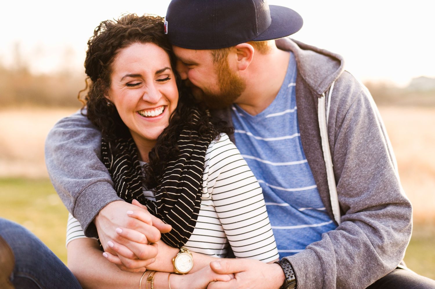 Warm sunset photo of a couple cuddling in a field, woman smiling while man kisses her cheek, dressed casually in fall layers.
