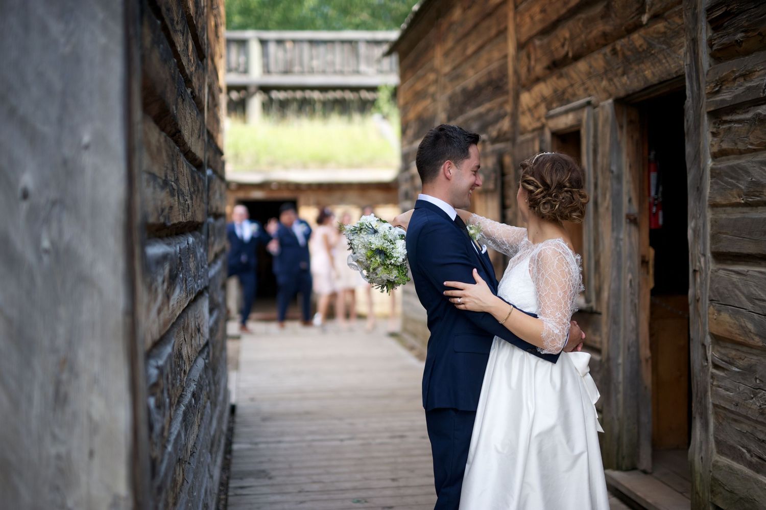 Bride touches groom’s lapel lovingly as guests gather in the background of a heritage village.