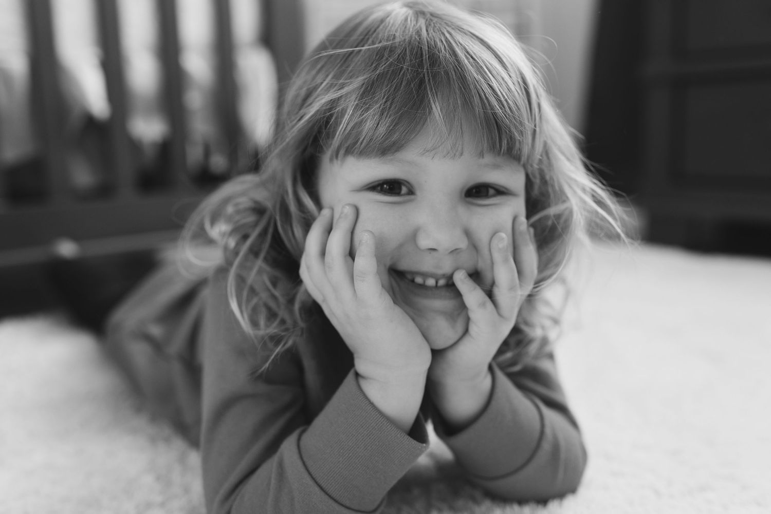 Young girl lying on her stomach on a porch, smiling into the camera with hands on her cheeks.