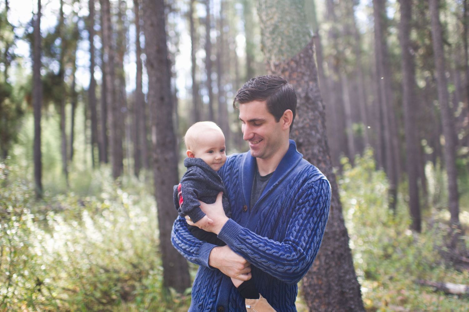 Dad smiles holding baby in forest light. Jasper National Park family photography with father and child.