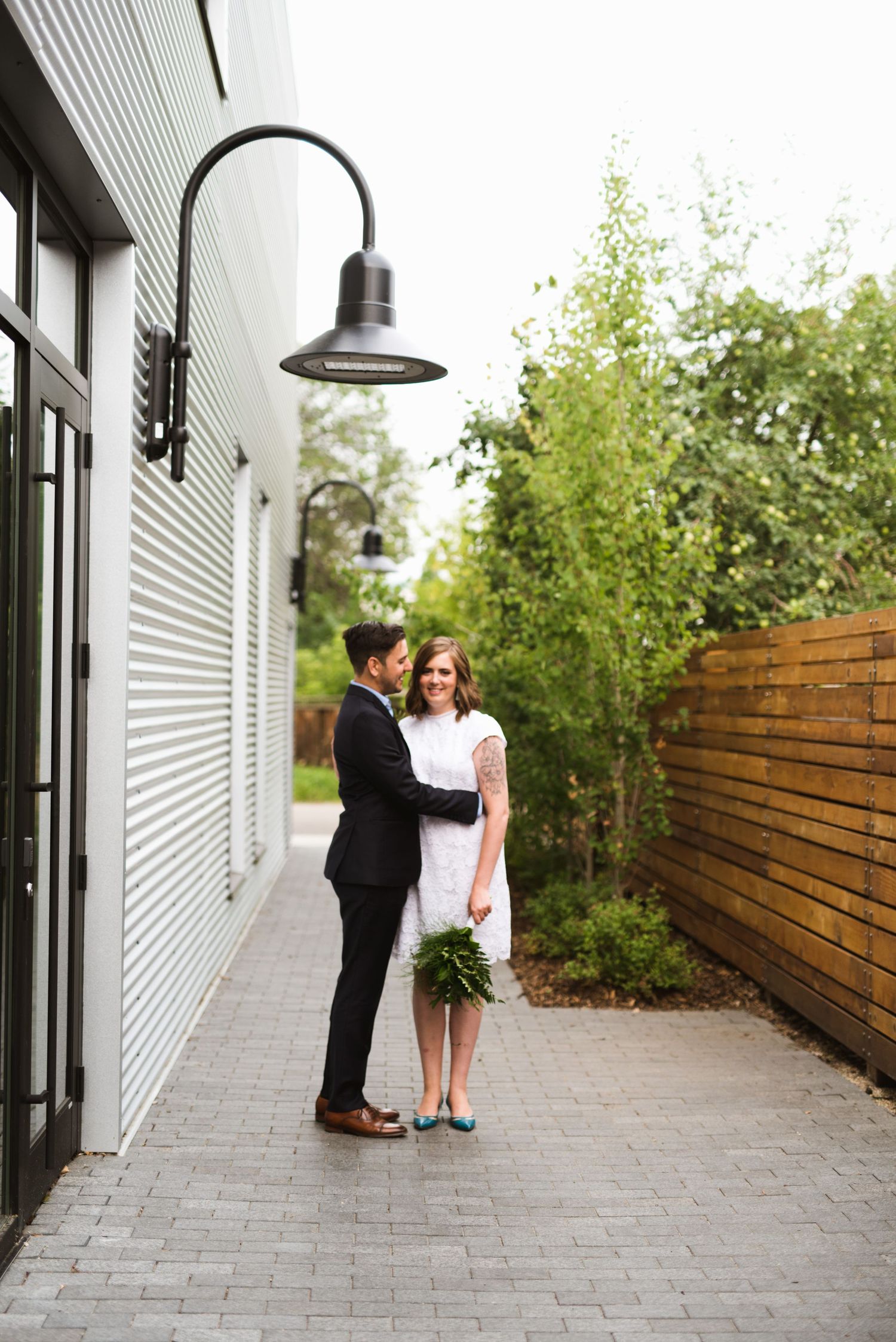 Urban elopement photo with bride and groom standing under black wall light in modern alleyway with bamboo and wood fencing.