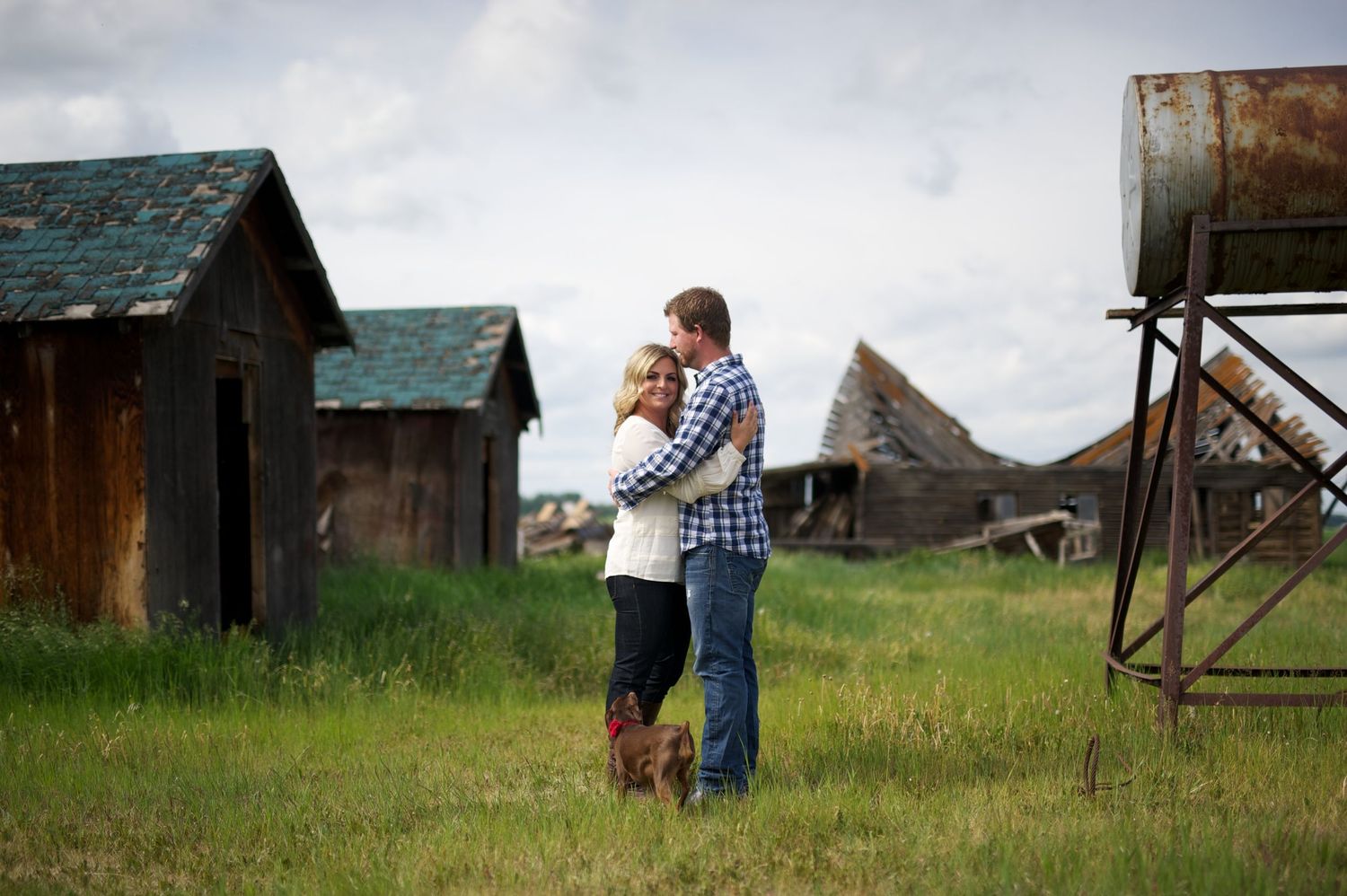 Couple hugging in a wide field surrounded by abandoned farm buildings and an old water tank, with moody skies and prairie energy.