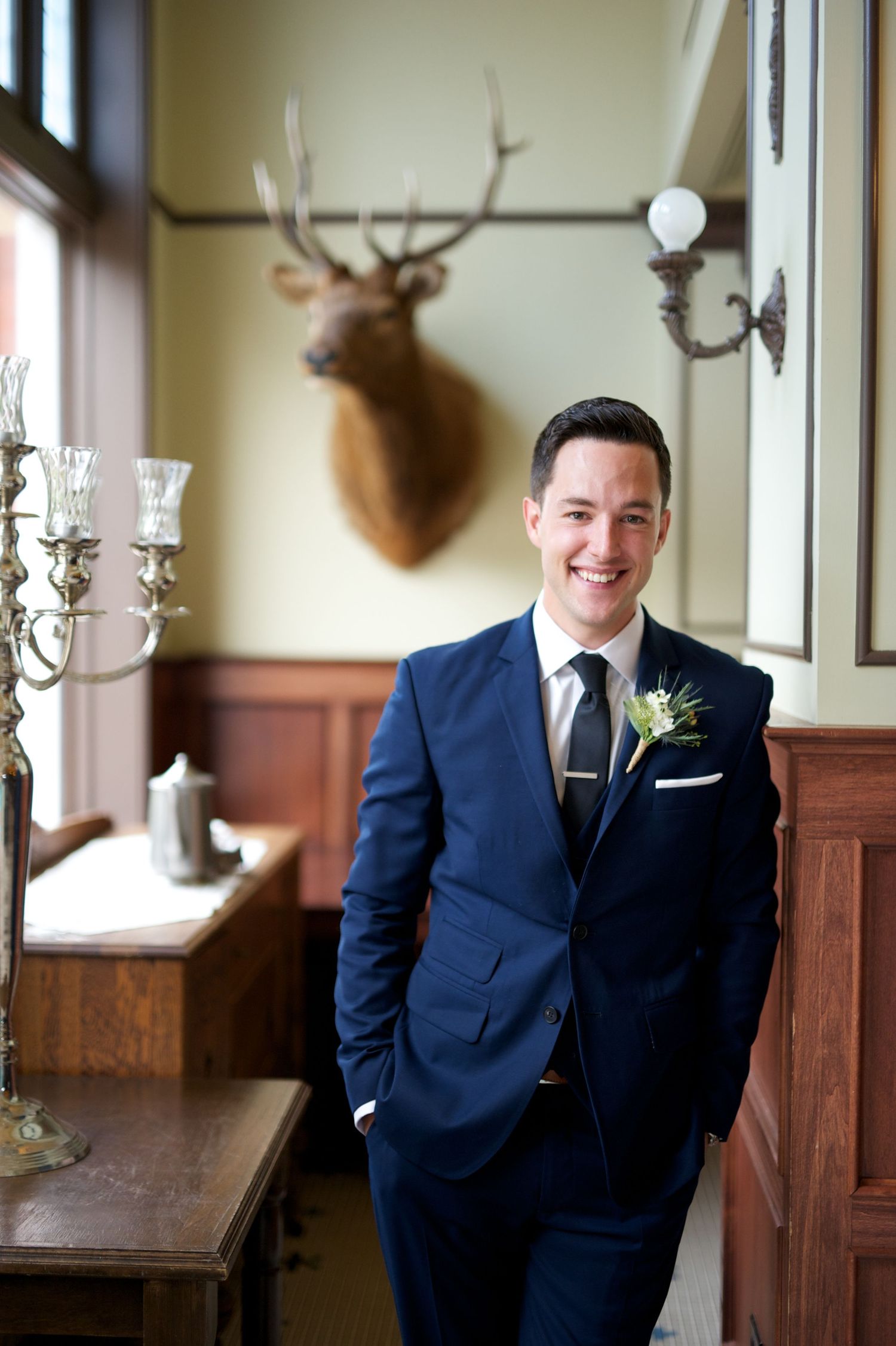Groom smiles in a vintage room with antlers on the wall and warm wood details. Classic and relaxed.
