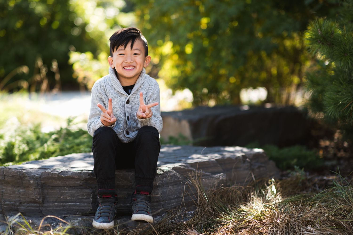 Smiling boy flashing double peace signs while sitting on a log in a sunny park setting.