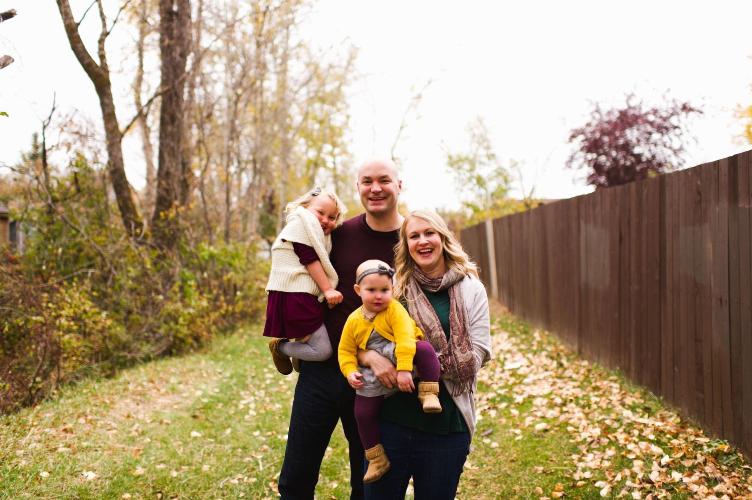 Family of four smiling in an autumn pathway with golden leaves, carrying two children
