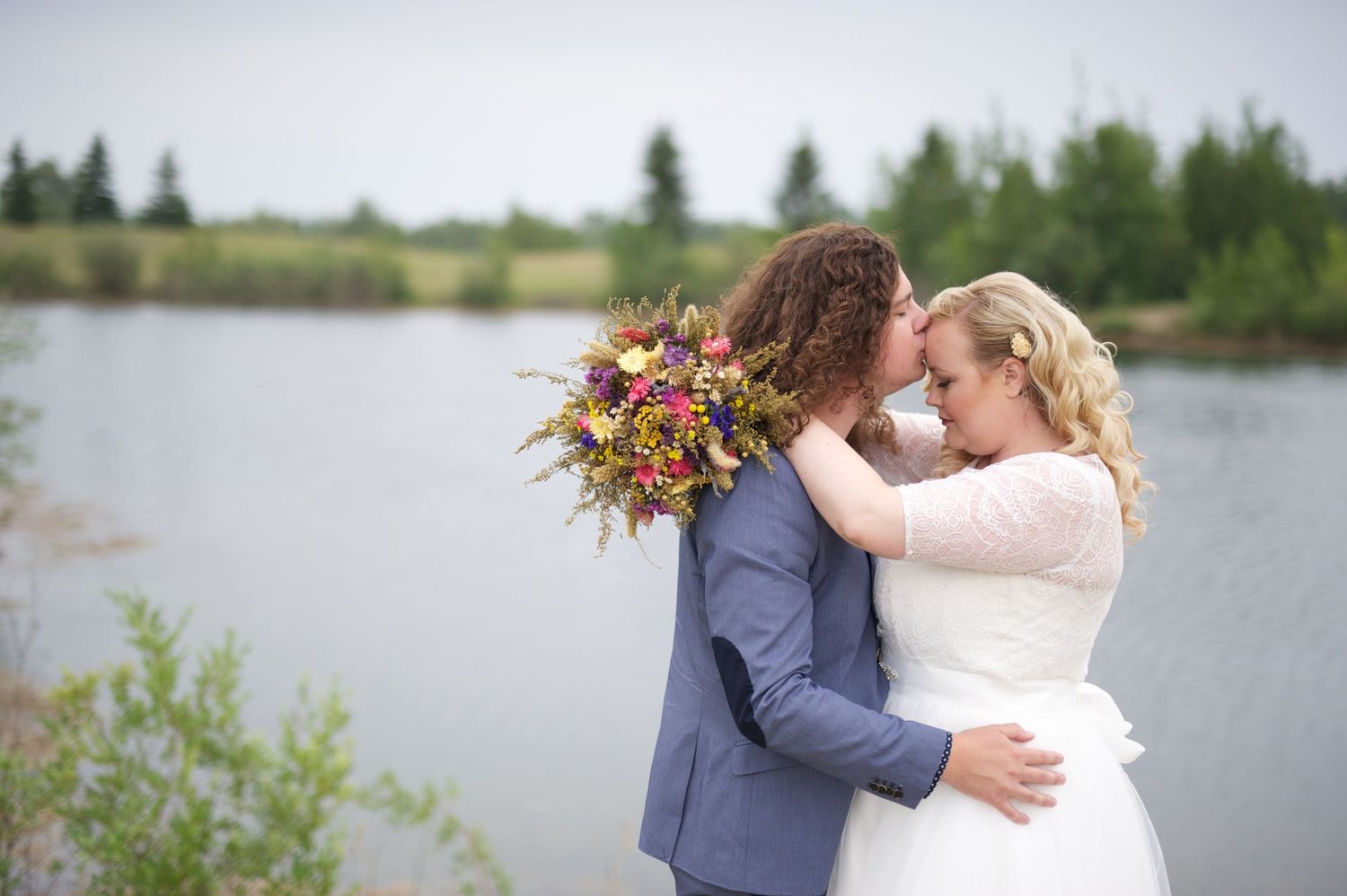 Couple sharing a kiss beside a calm pond, bride’s arms wrapped around groom with wildflower bouquet in focus.