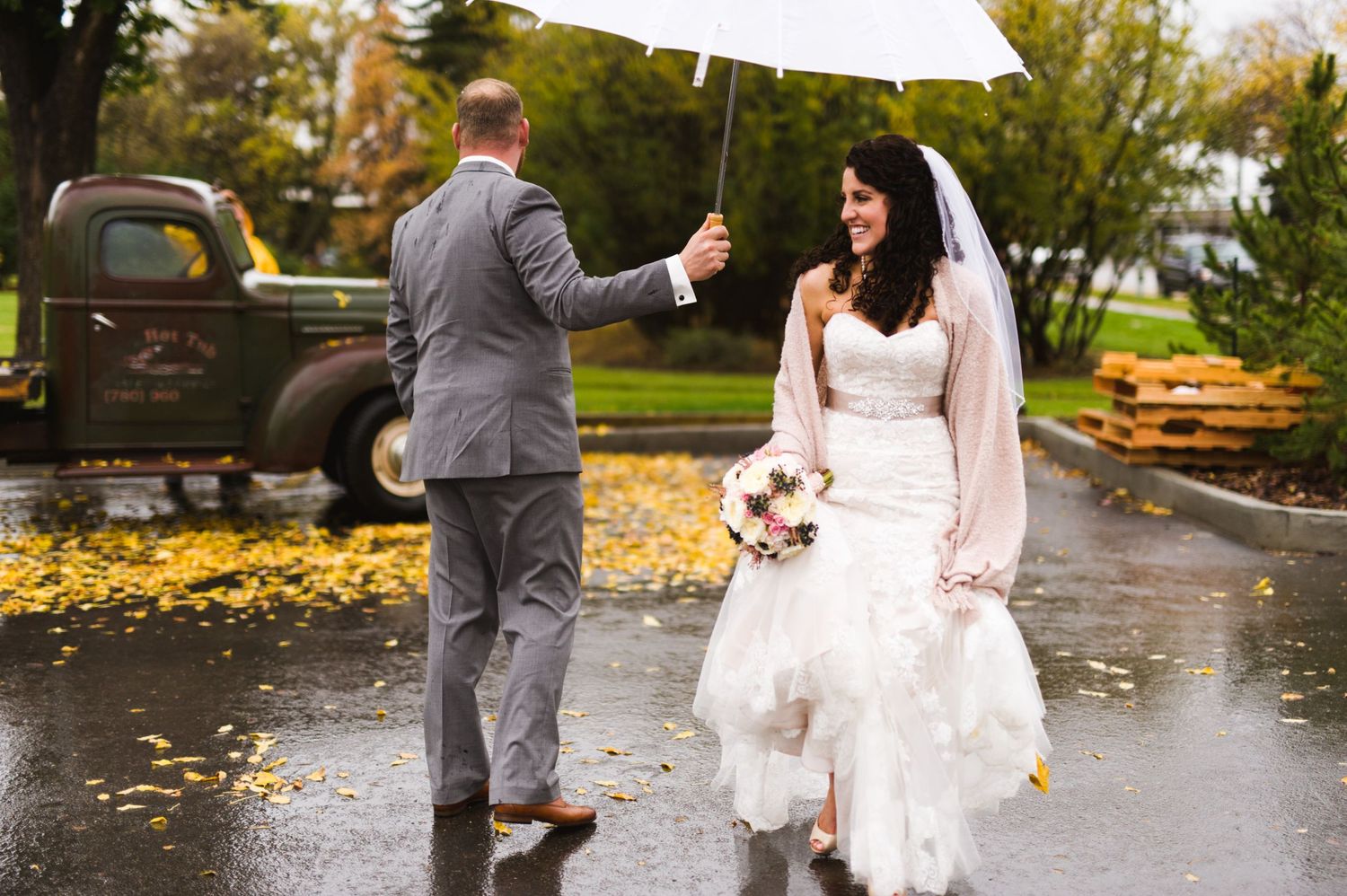 Groom holding a white umbrella for his bride as they walk across a wet pavement in front of a vintage truck, bouquet in hand and raindrops o