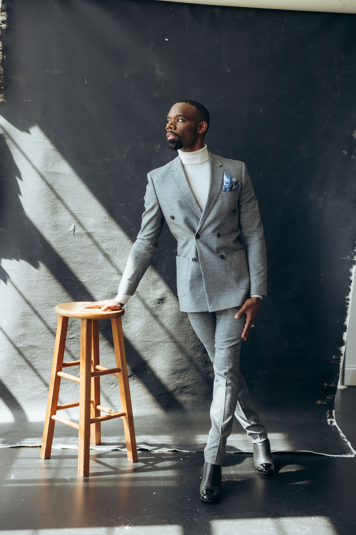 Fashion model in grey suit poses with wooden stool against dark backdrop with dramatic shadows.
