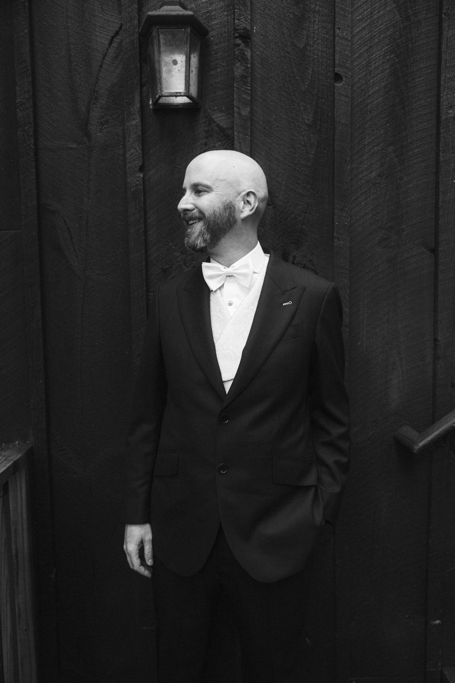 Groomsmen in formal black tuxedos standing against dark wooden wall.