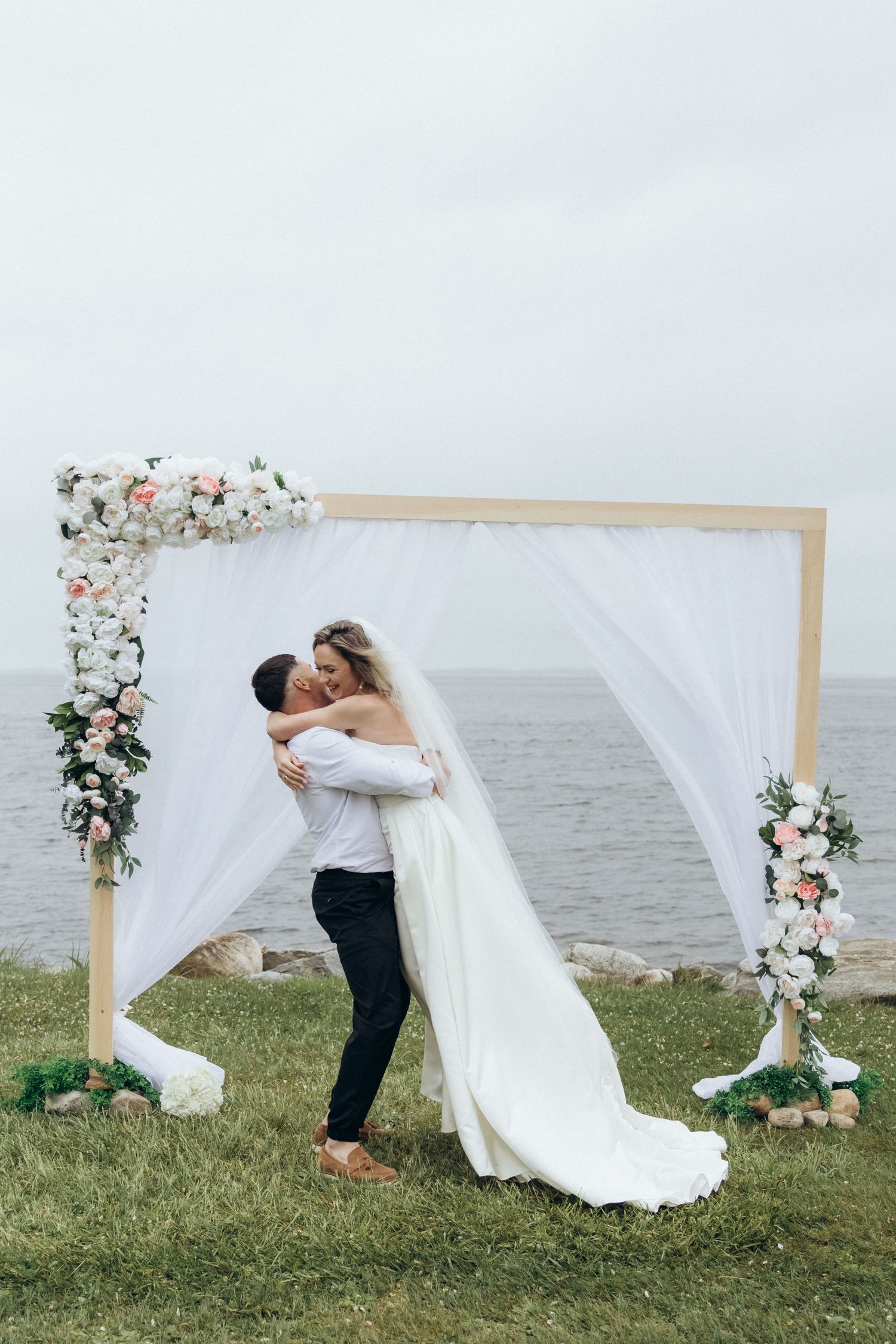 Wedding ceremony setup on a waterfront with white fabric arch and floral decorations.
