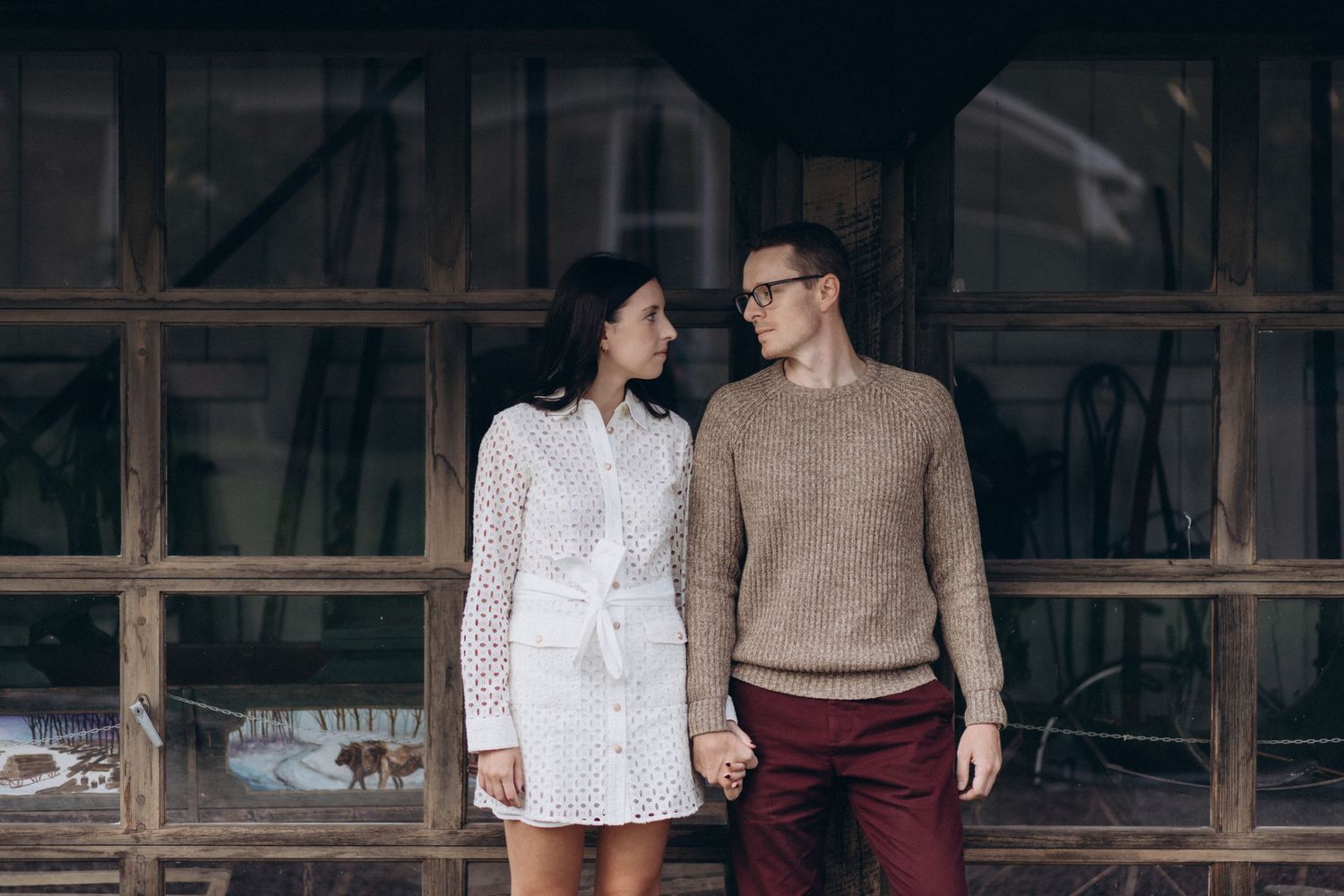 A couple stands together in front of a rustic wooden building wearing complementary autumn outfits.