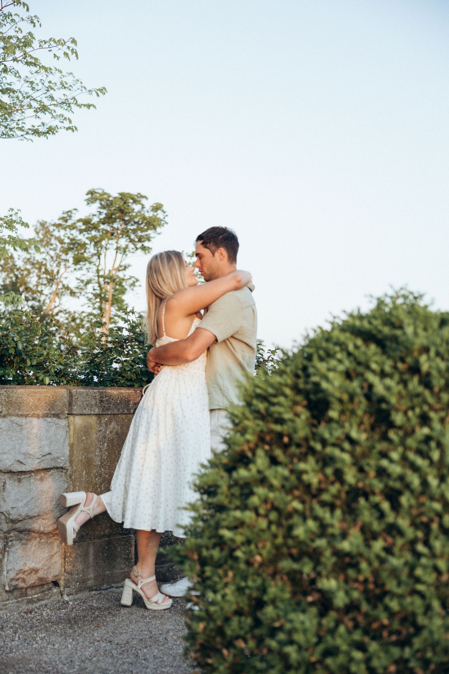 A couple sitting together along a stone wall with greenery in a romantic outdoor setting.