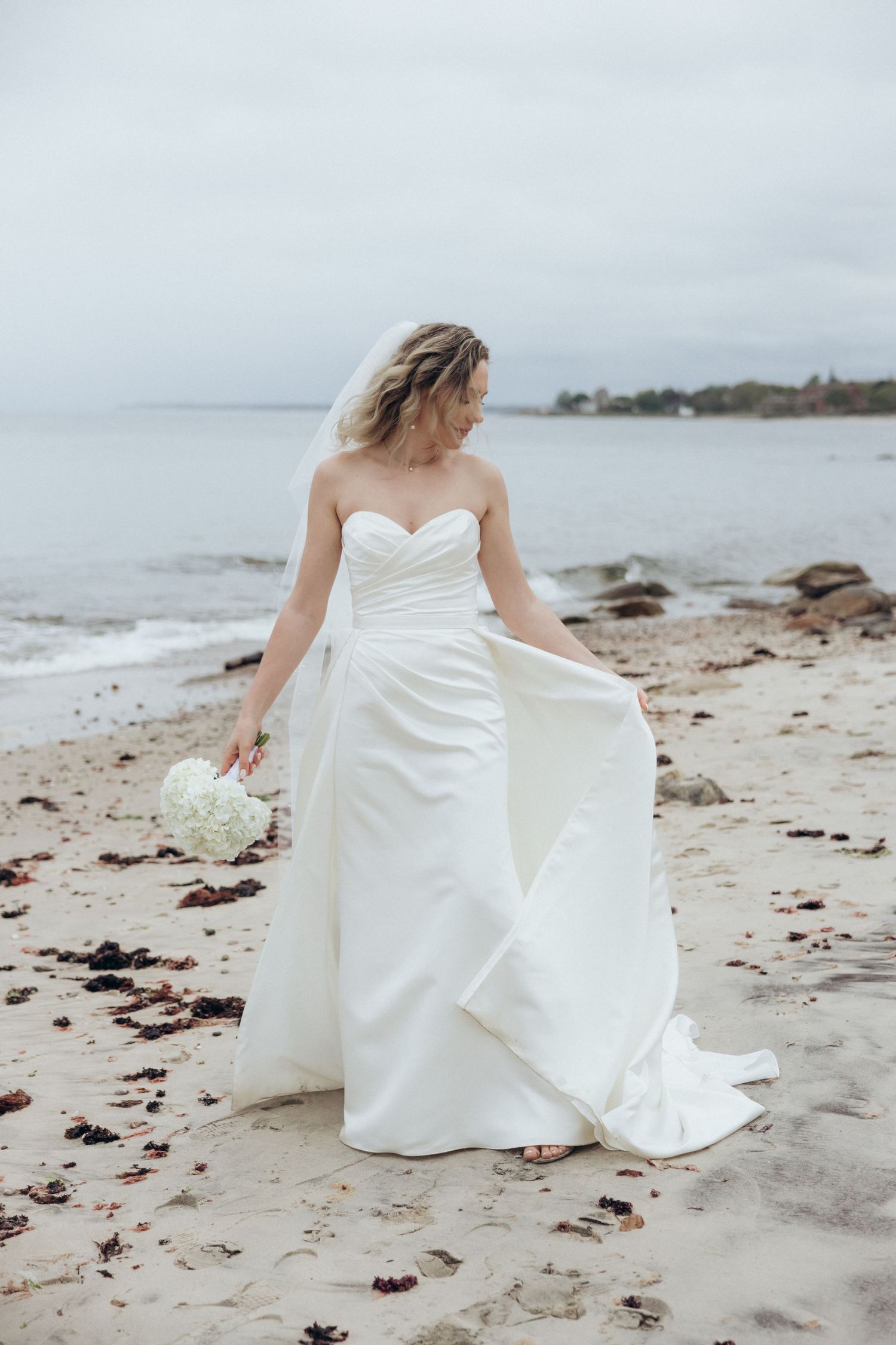 White wedding dress flowing in the breeze on a sandy beach with seaweed.