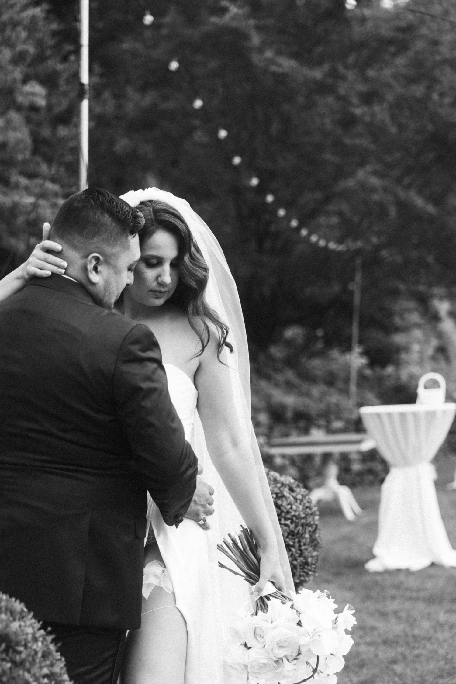 Tender black and white wedding photo captured outdoors with string lights in background.
