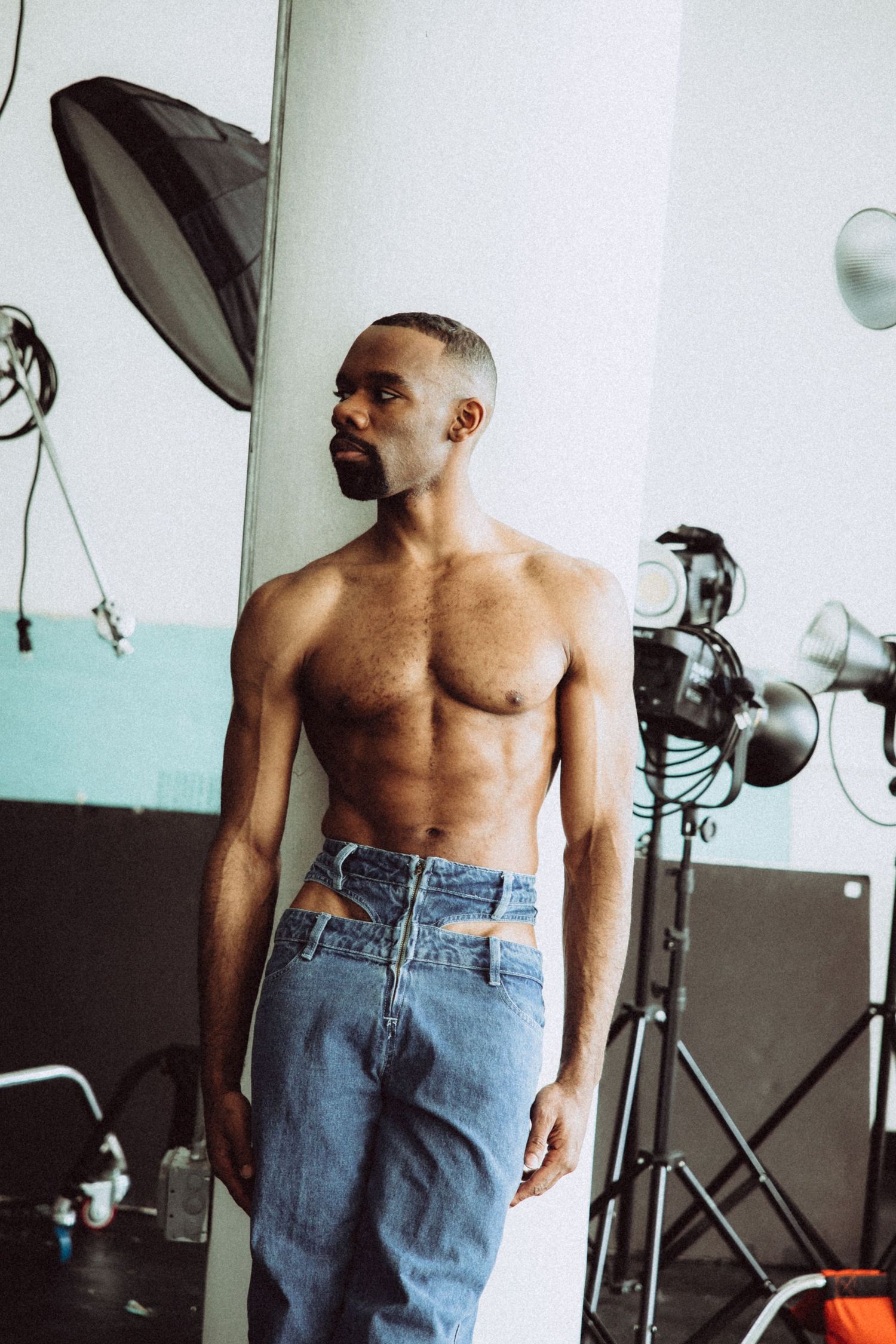 Shirtless model in high-waisted jeans poses in studio with lighting equipment.