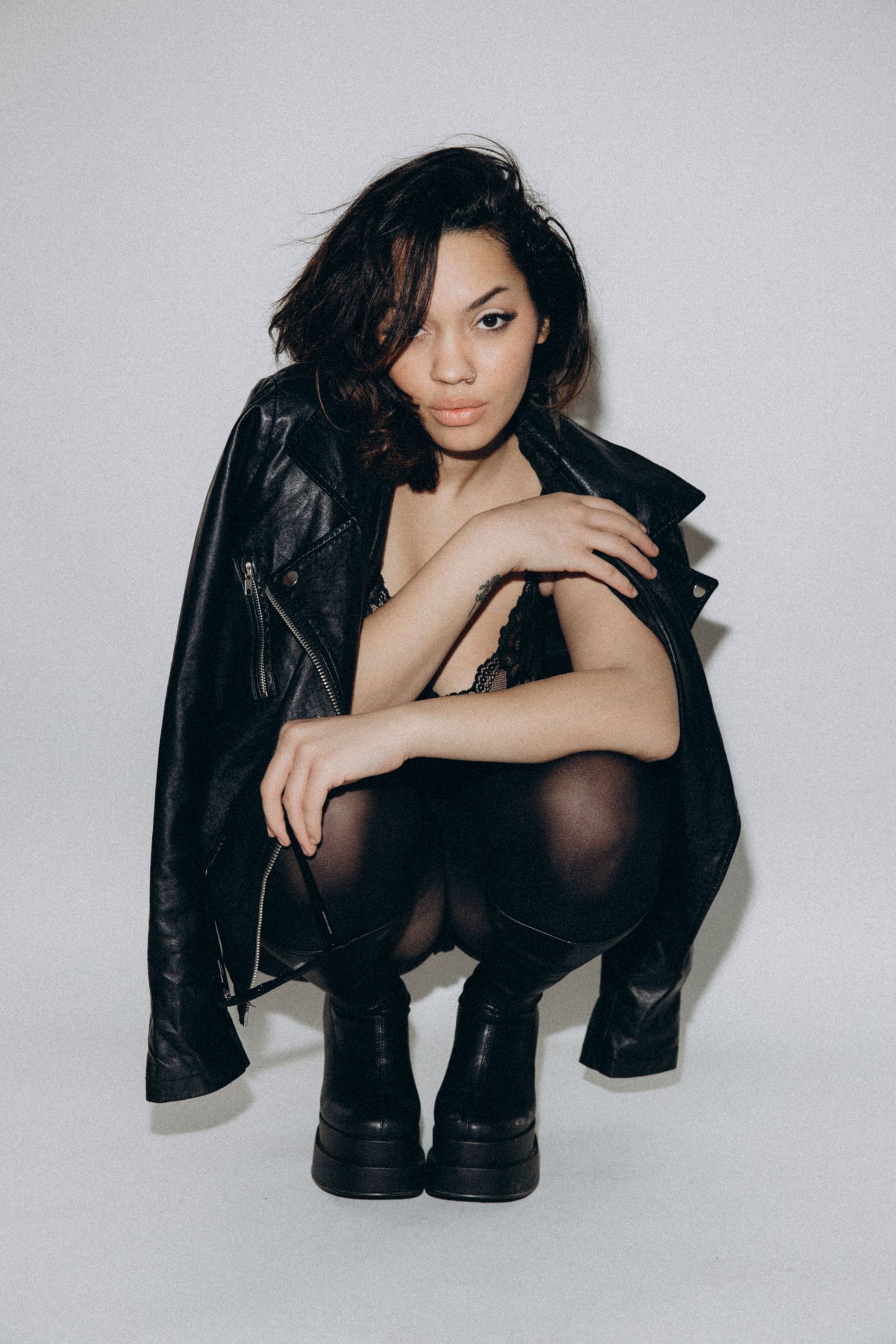 Model in black leather jacket crouches in edgy pose against white studio background.