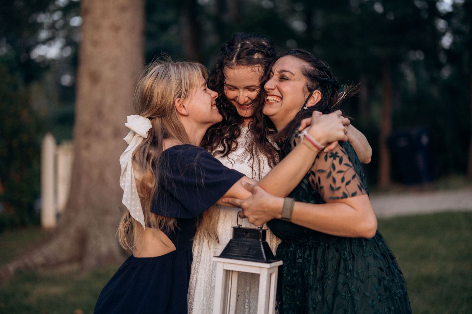 Three wedding guests share emotional embrace and laughter during outdoor celebration.