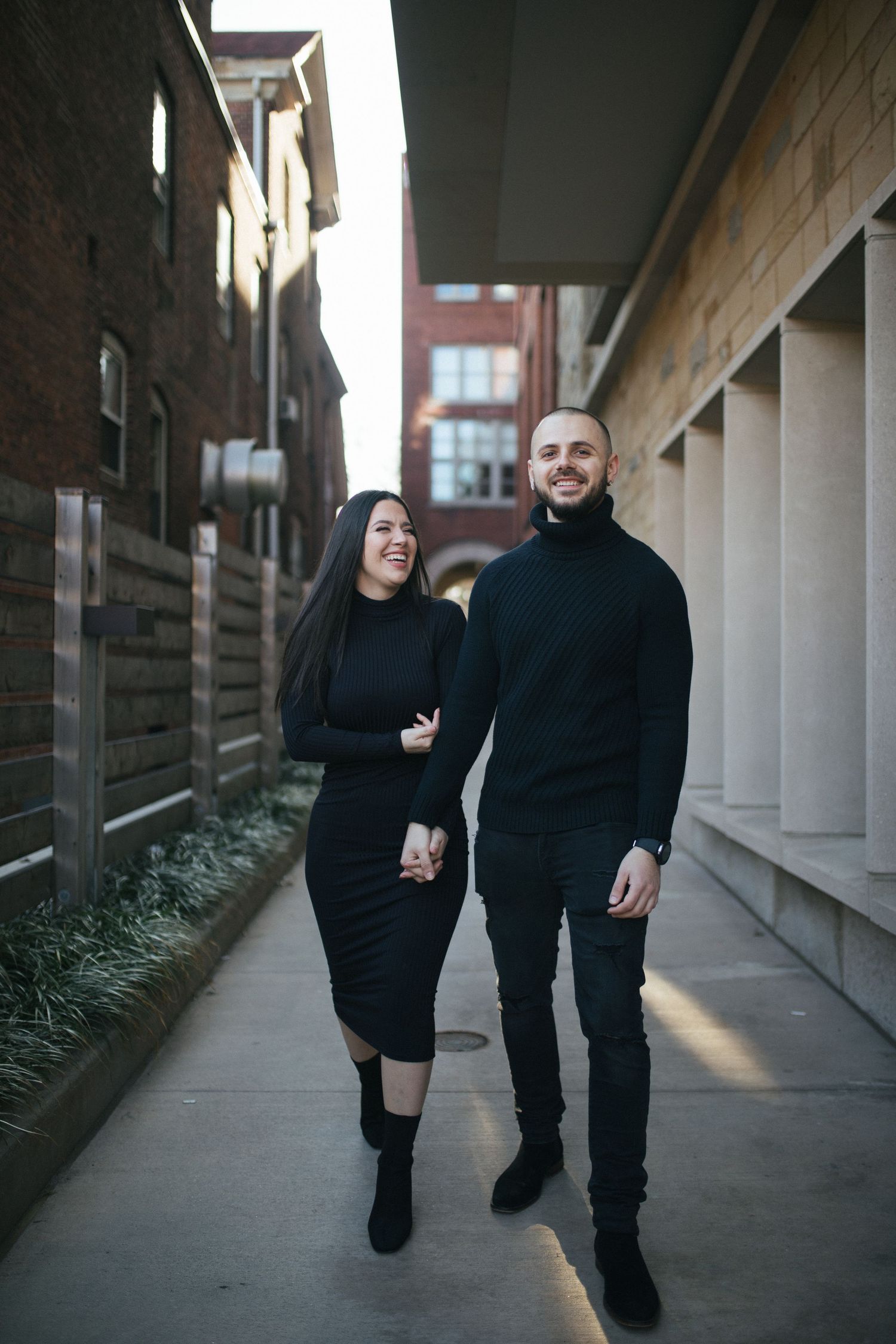 Couple in matching black outfits walking together down a covered walkway.