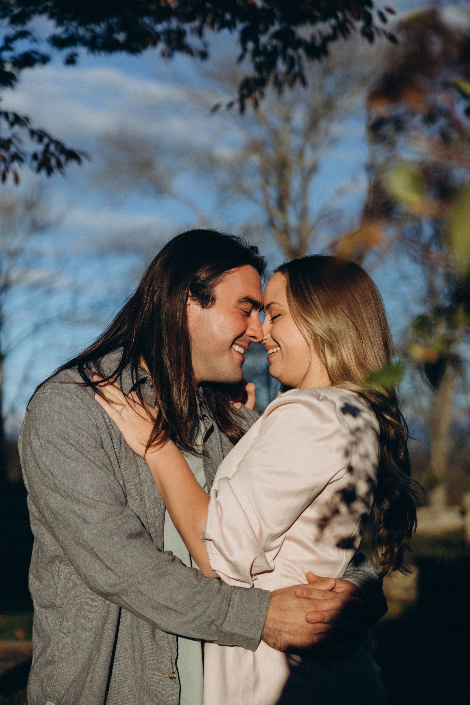 A couple sharing a tender kiss outdoors during golden hour with trees in the background.