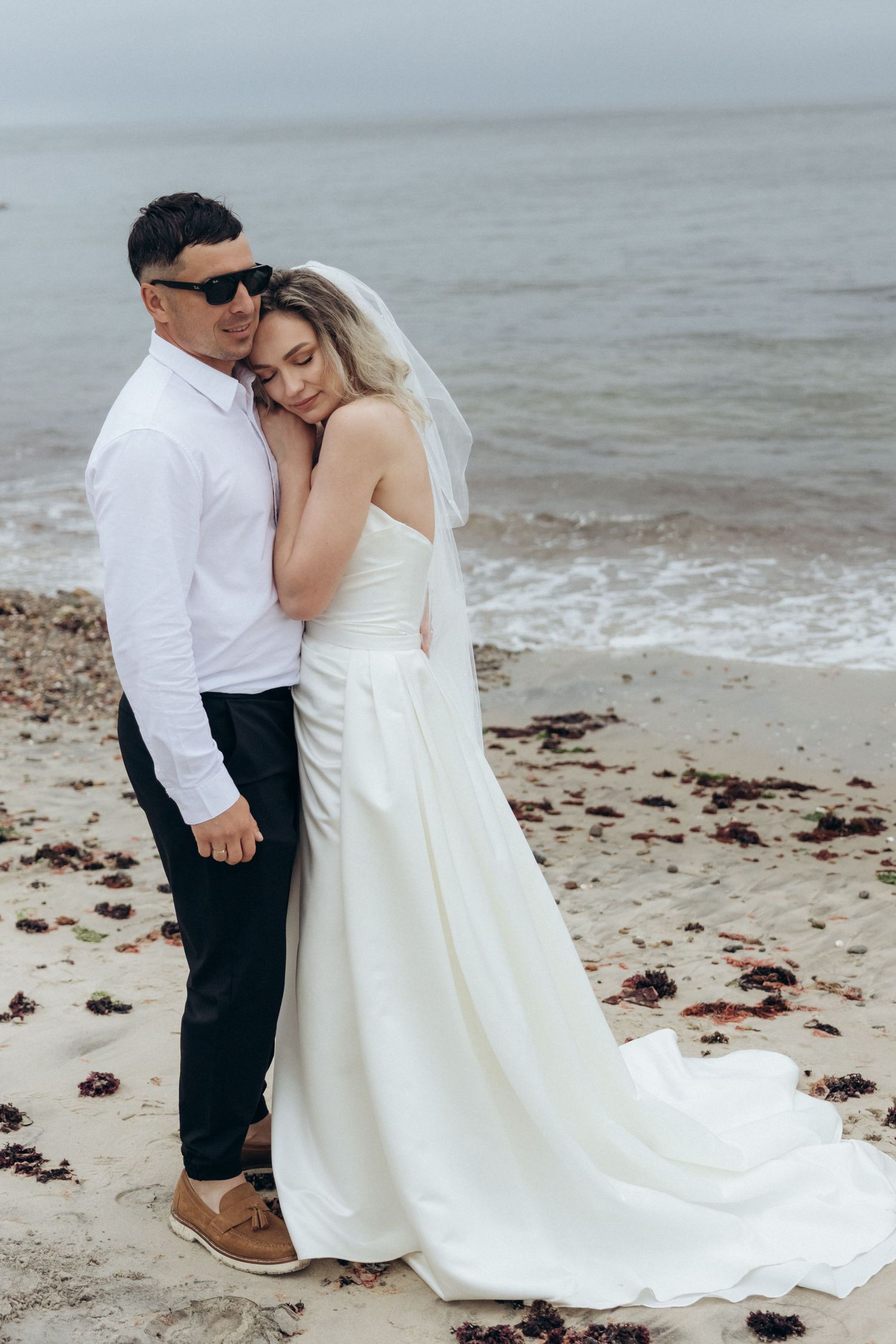 Couple in wedding attire embracing on a scenic beach with waves in the background.