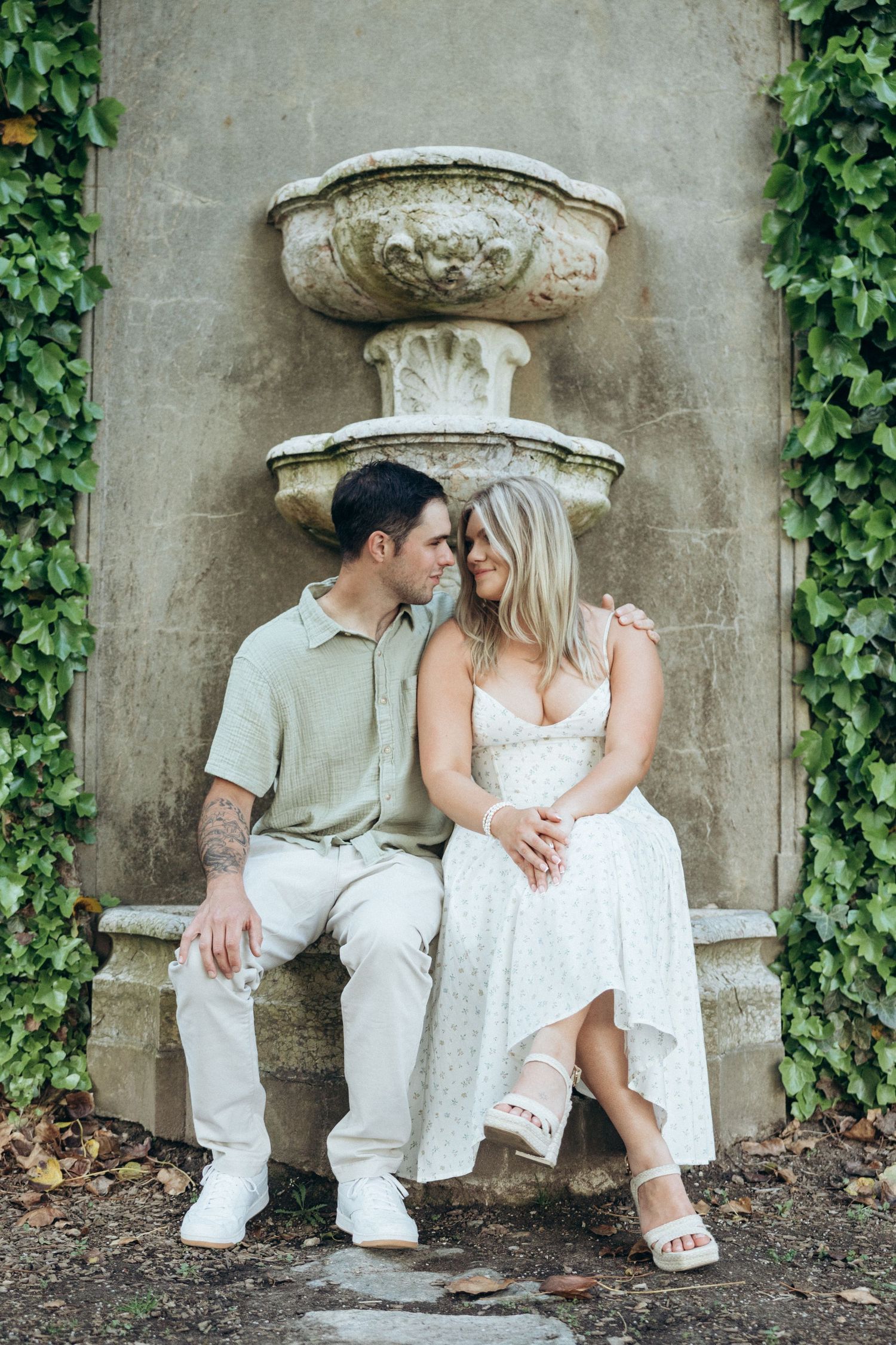 A couple in white outfits sitting together against a stone wall covered with ivy.