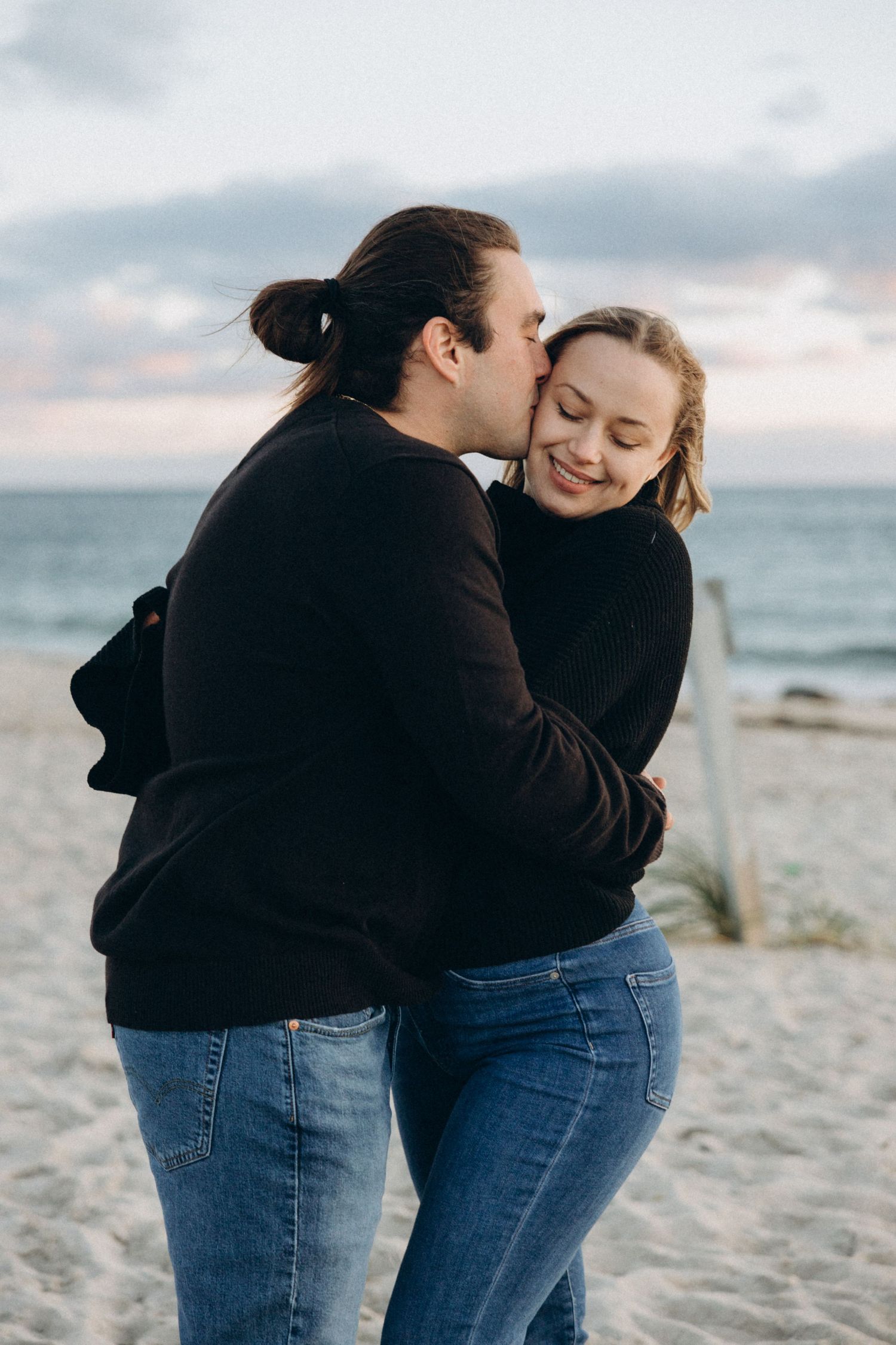 Couple in matching black hoodies and jeans sharing intimate moments on a beach at sunset.