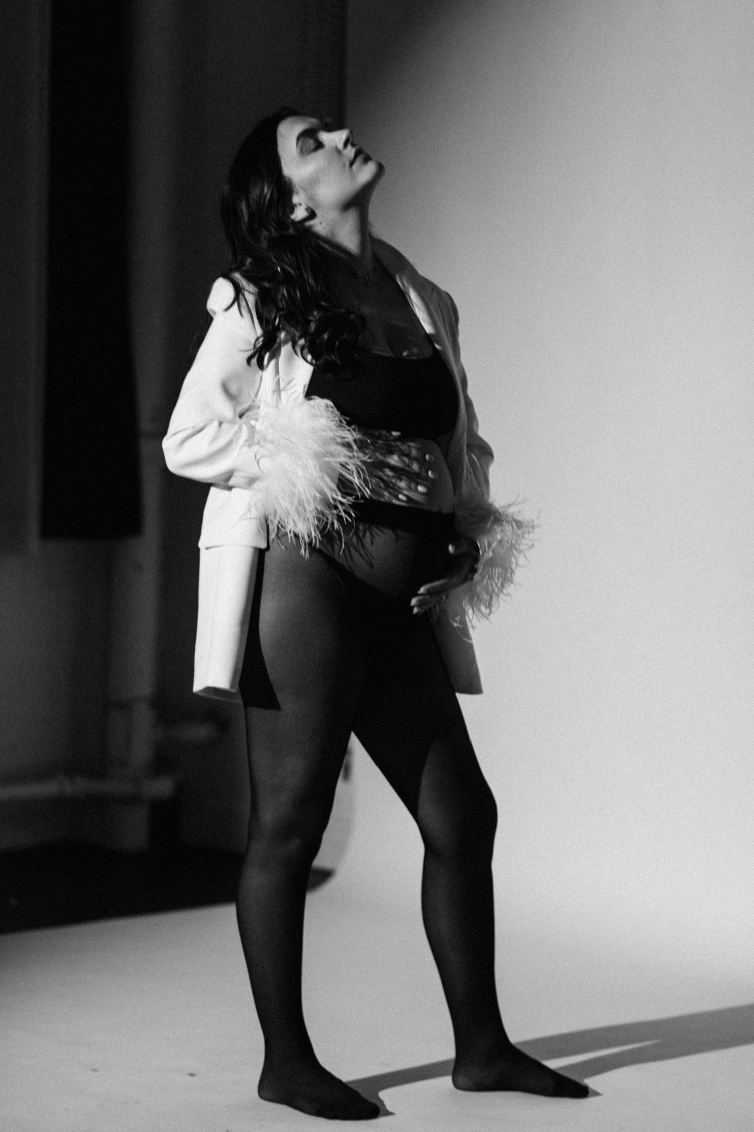 Dramatic fashion pose in black bodysuit and white jacket with feather details.