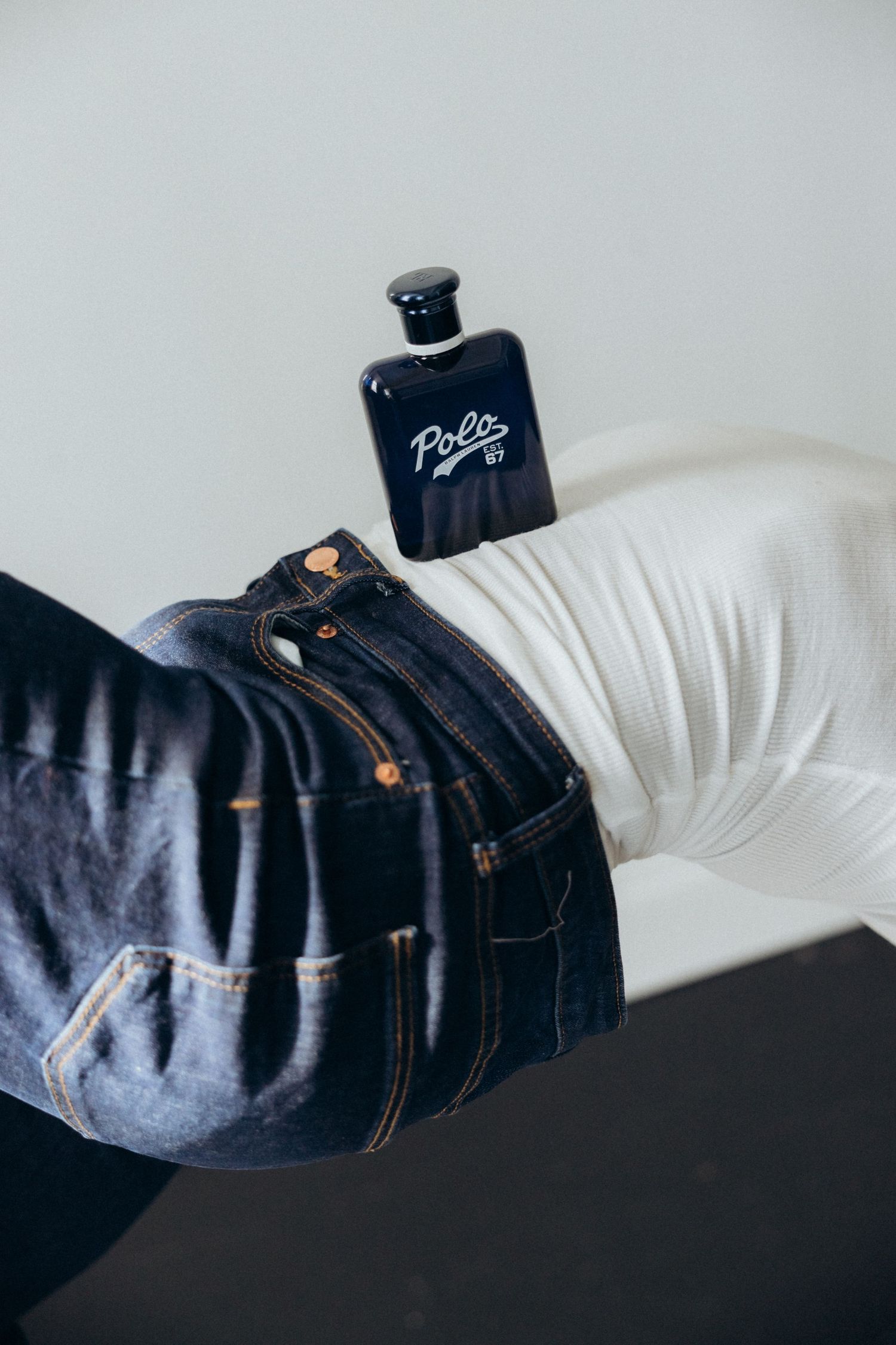 Dark blue perfume bottle rests on denim jeans against white background.