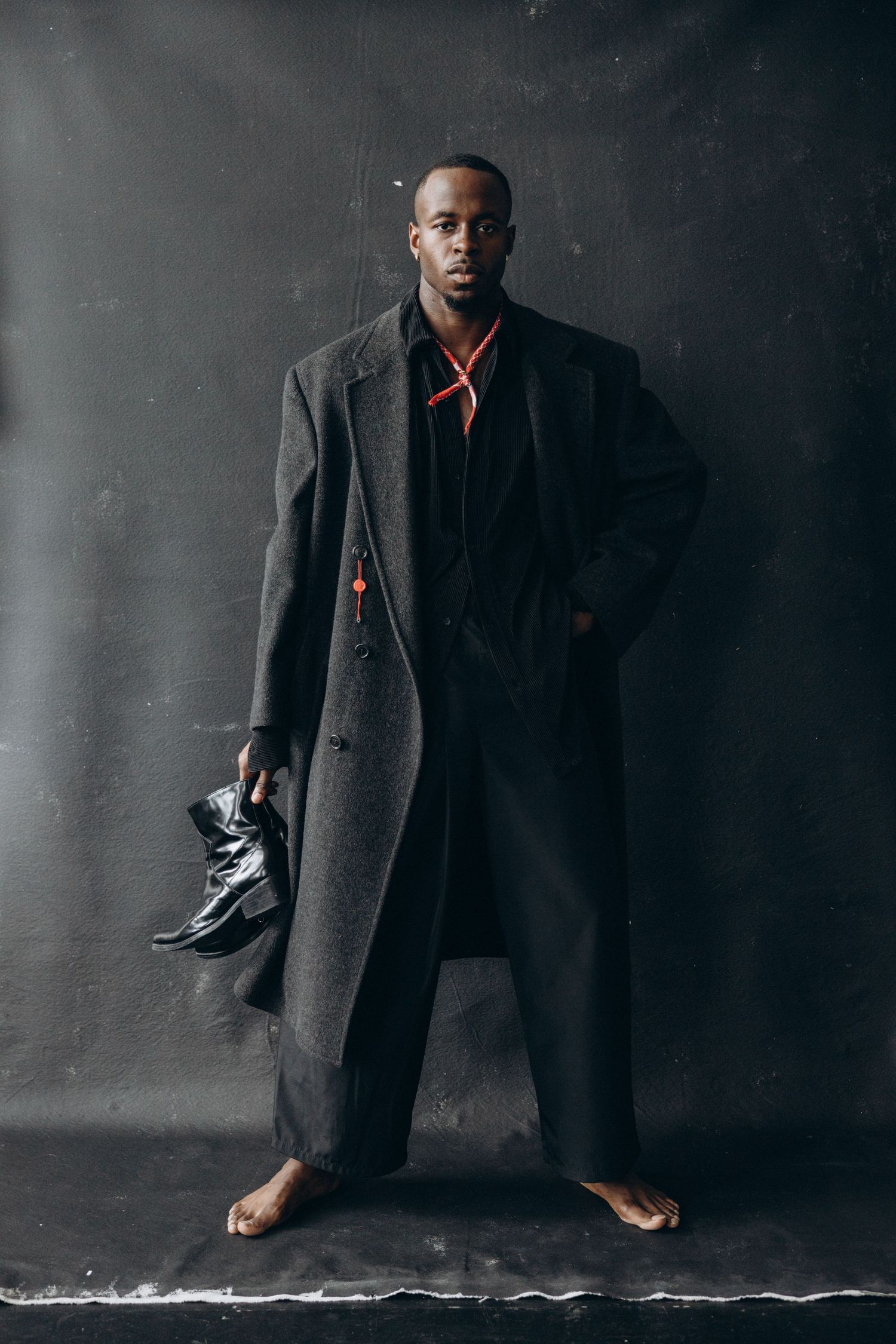 Model wearing long gray coat holds shoes in artistic monochromatic studio portrait.