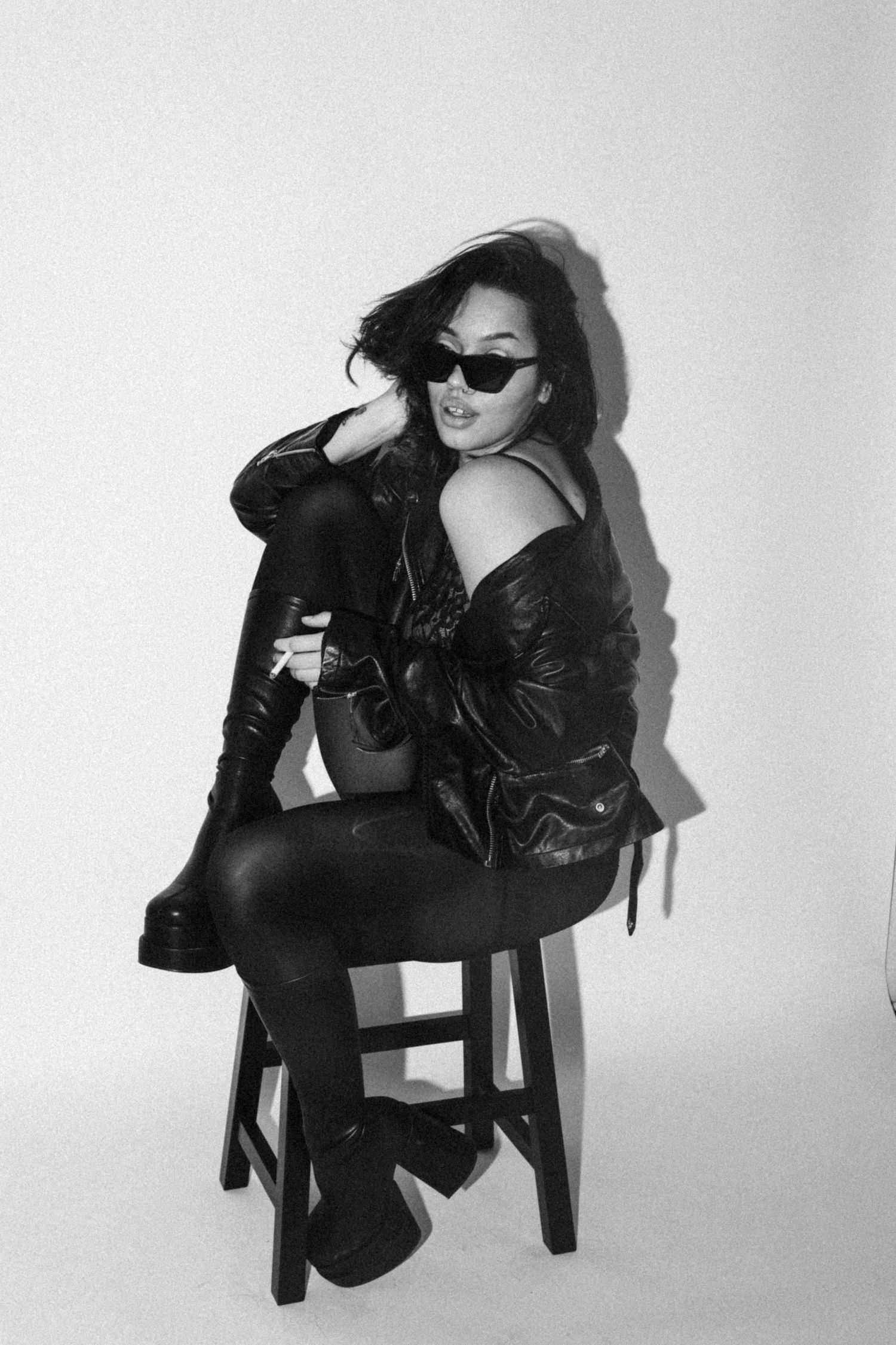 Fashion model poses on stool wearing sunglasses, leather jacket and combat boots in black and white photo.