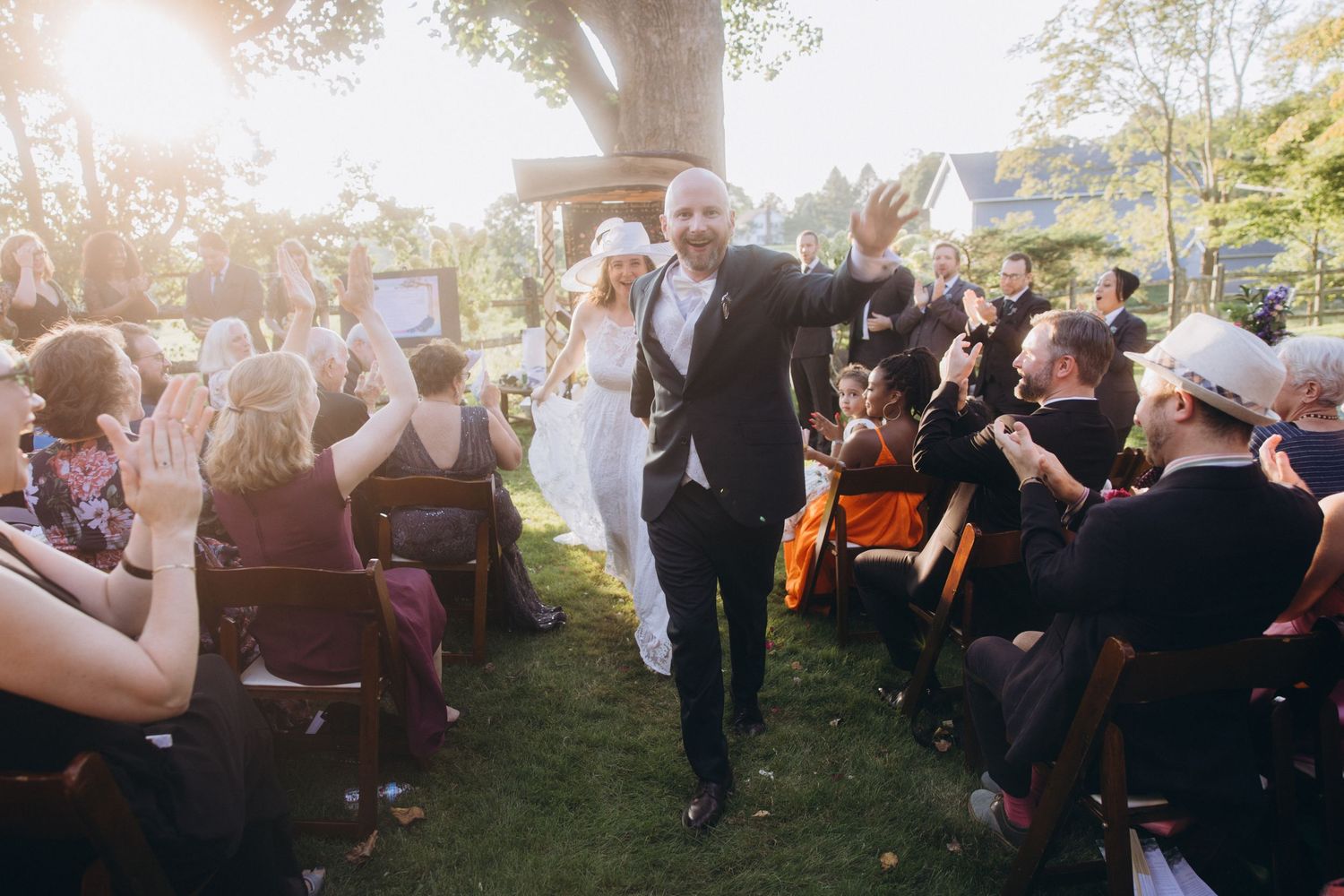 Lively outdoor wedding celebration with guests clapping and cheering in natural sunlight.