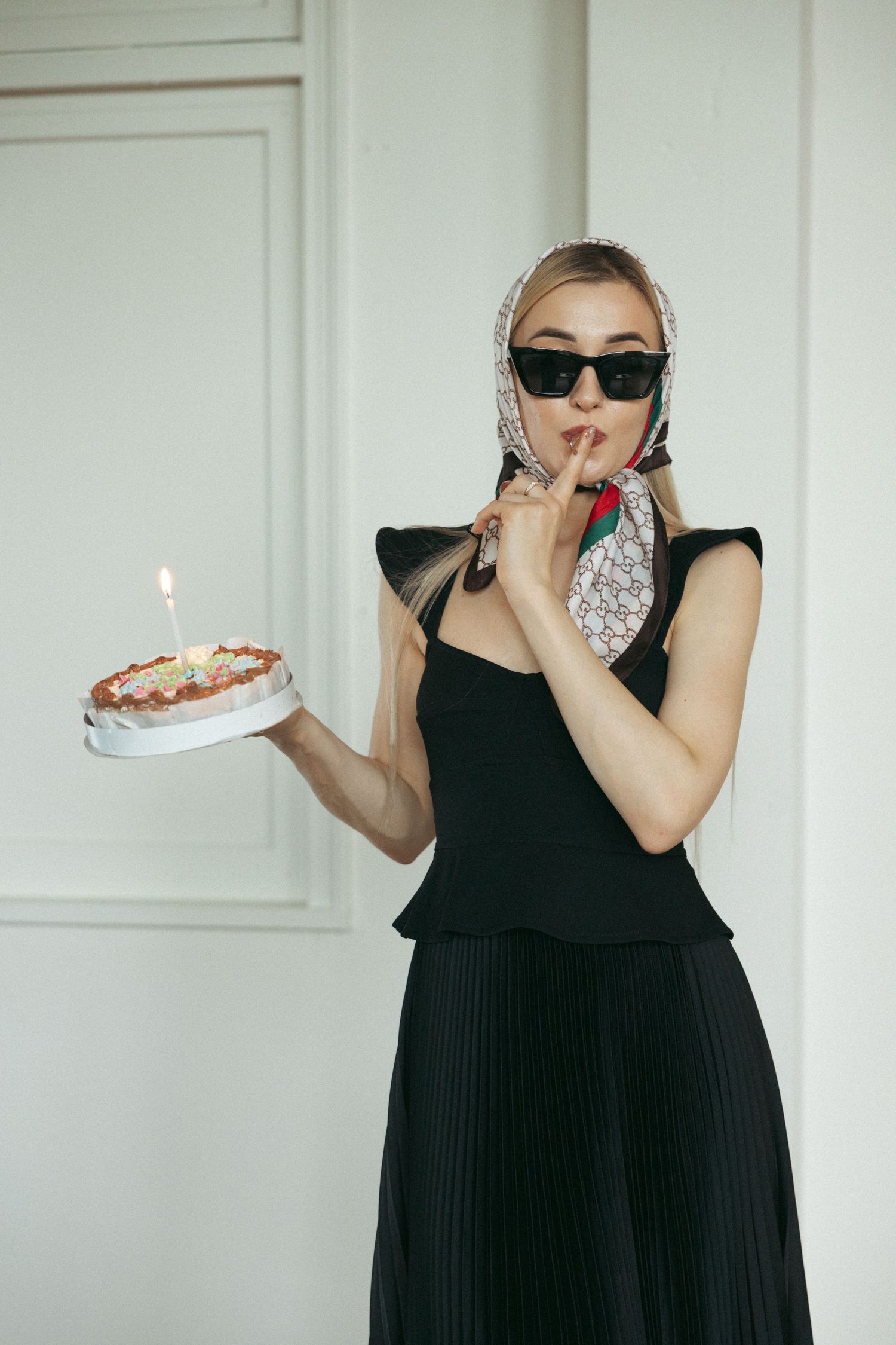 Fashion photoshoot featuring a woman in a black dress holding a birthday cake and wearing sunglasses.