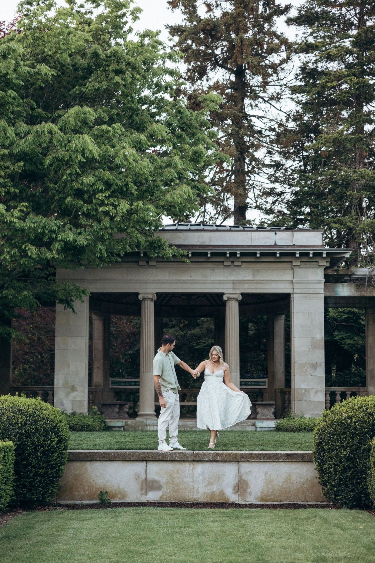 A couple in white clothing standing on a modern architectural terrace surrounded by manicured landscaping.