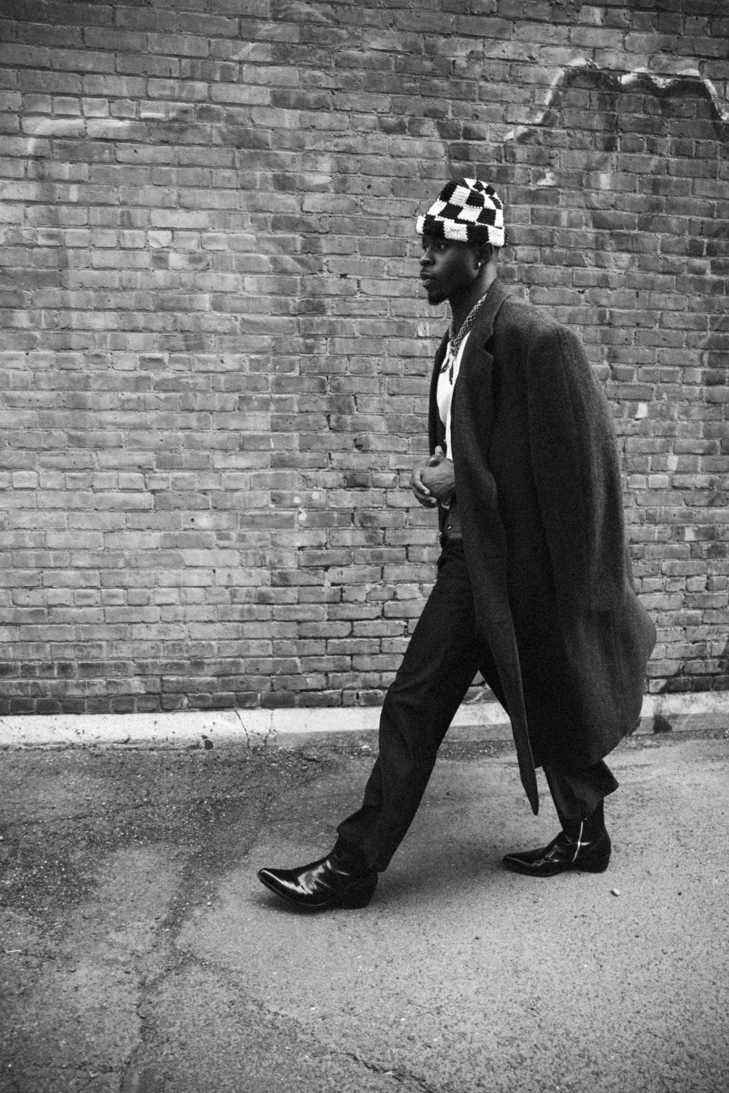 Black and white portrait of model in coat and checkered hat walking past brick wall.