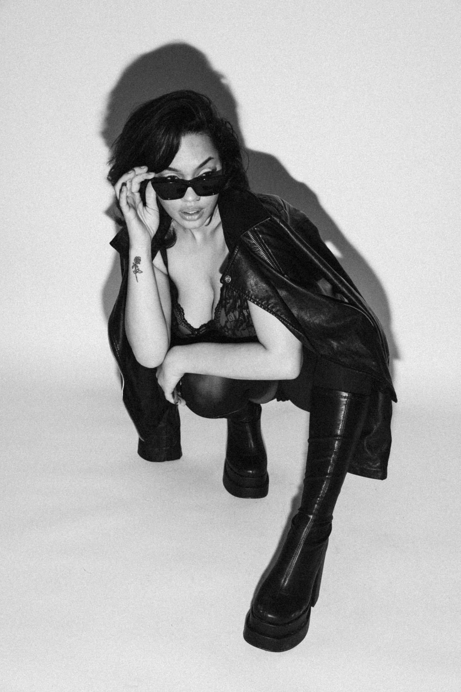 Fashion model in sunglasses and leather jacket poses in black and white studio shot.