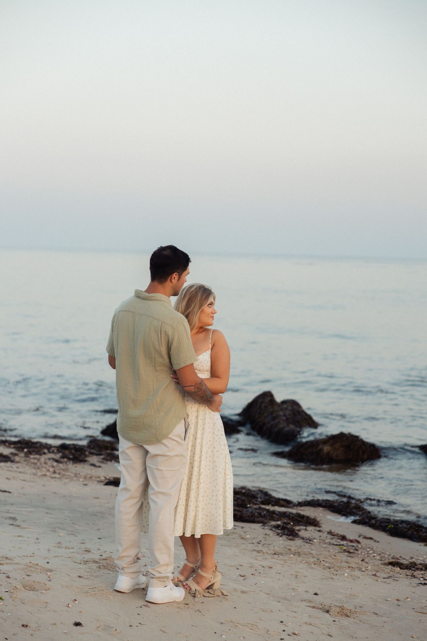 A couple sharing an intimate moment on a rocky beach at sunset with ocean waves in the background.