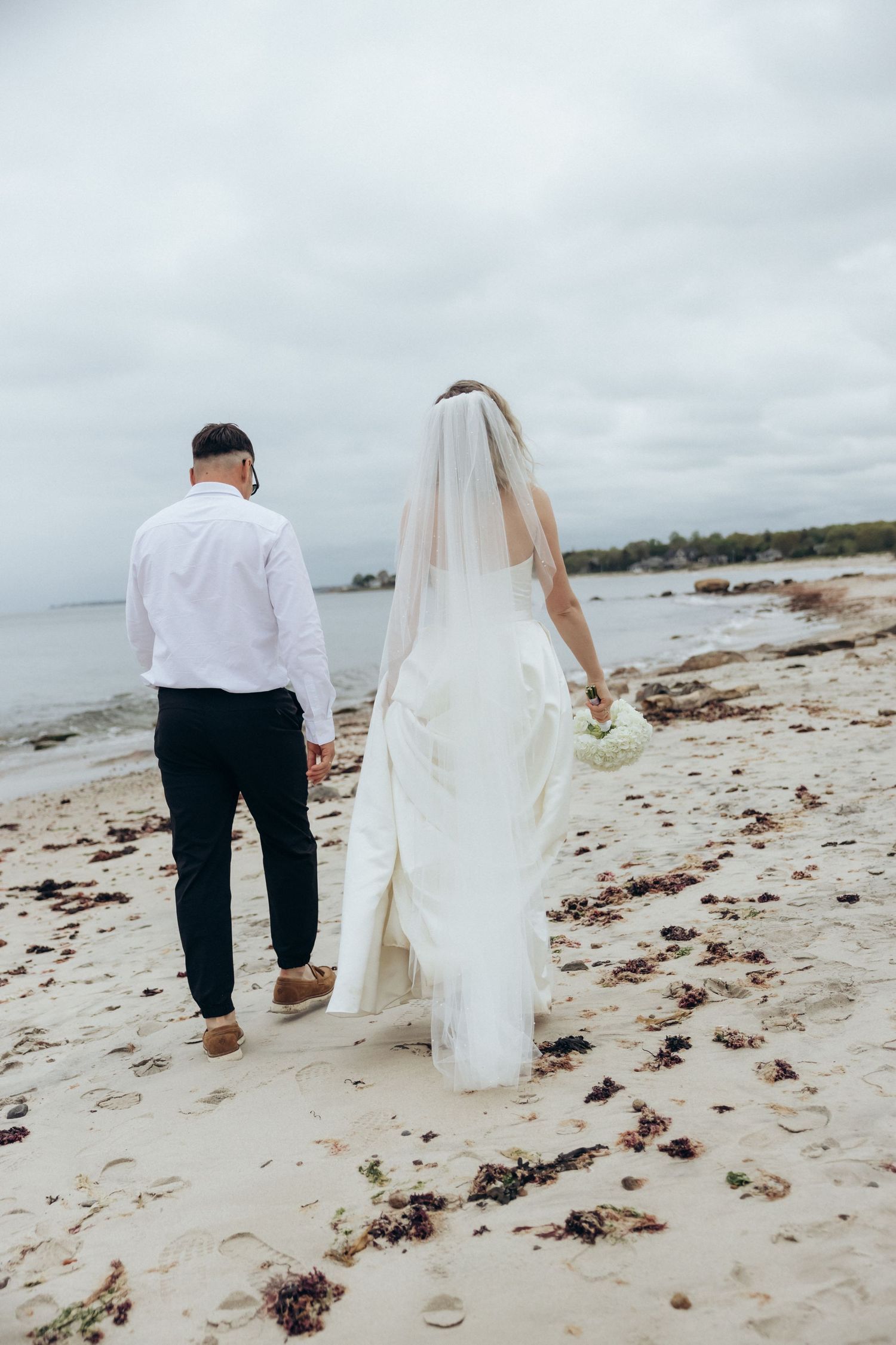 Wedding couple walking hand in hand along a seaweed-strewn beach at dusk.