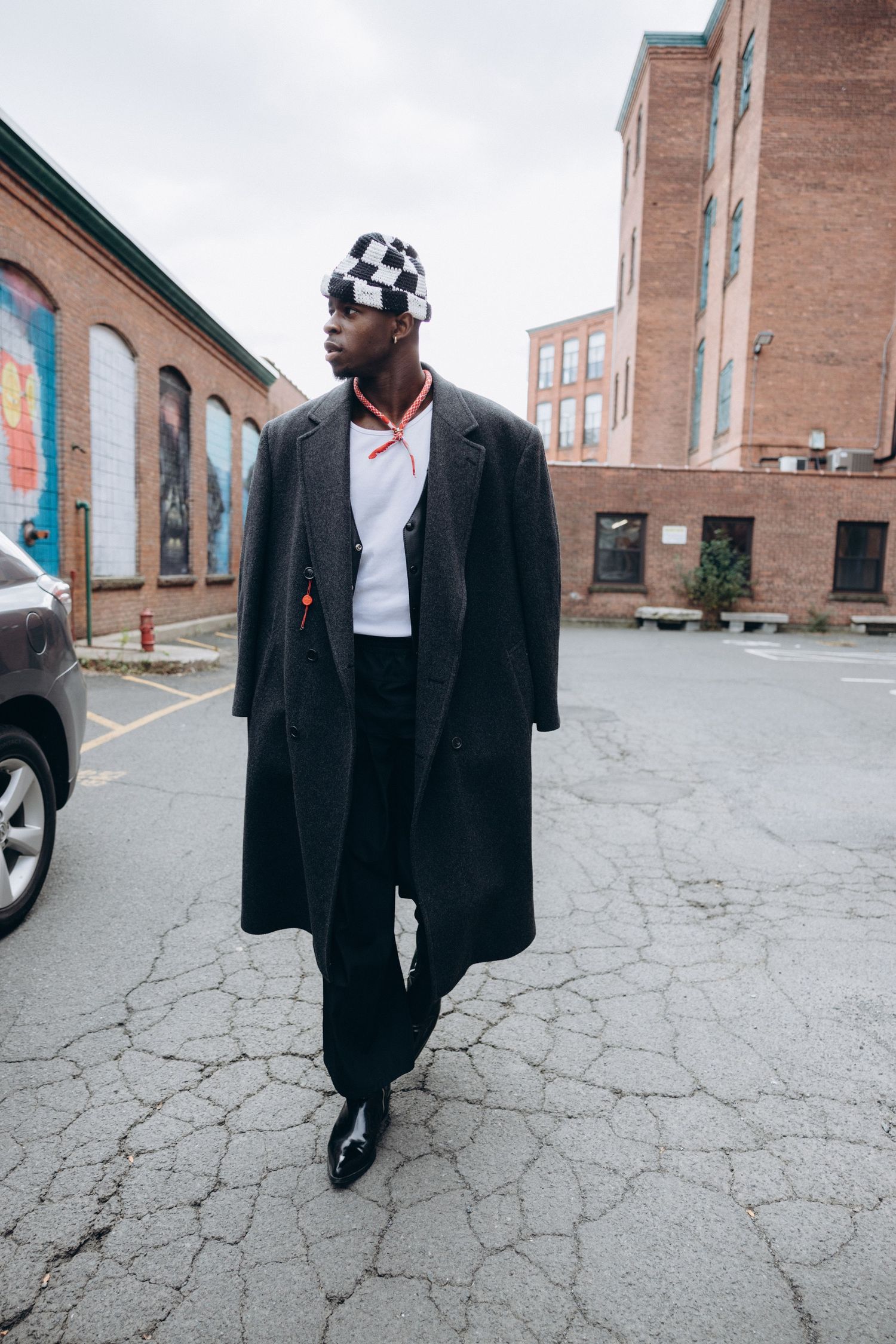 Street style photo of model in checkered headwear and long coat walking in urban setting.
