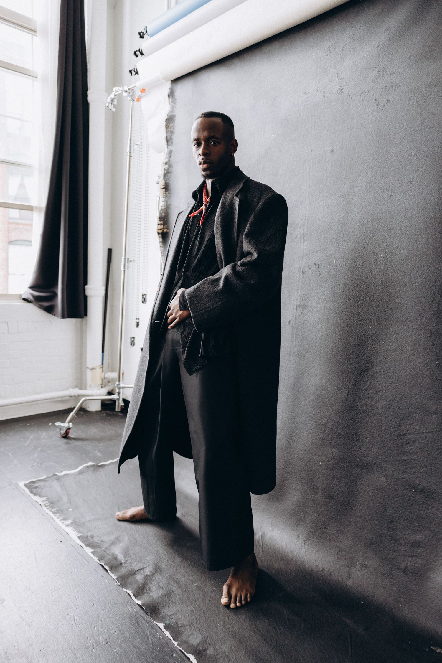 Fashion model in long black coat poses barefoot against studio wall in minimalist setting.