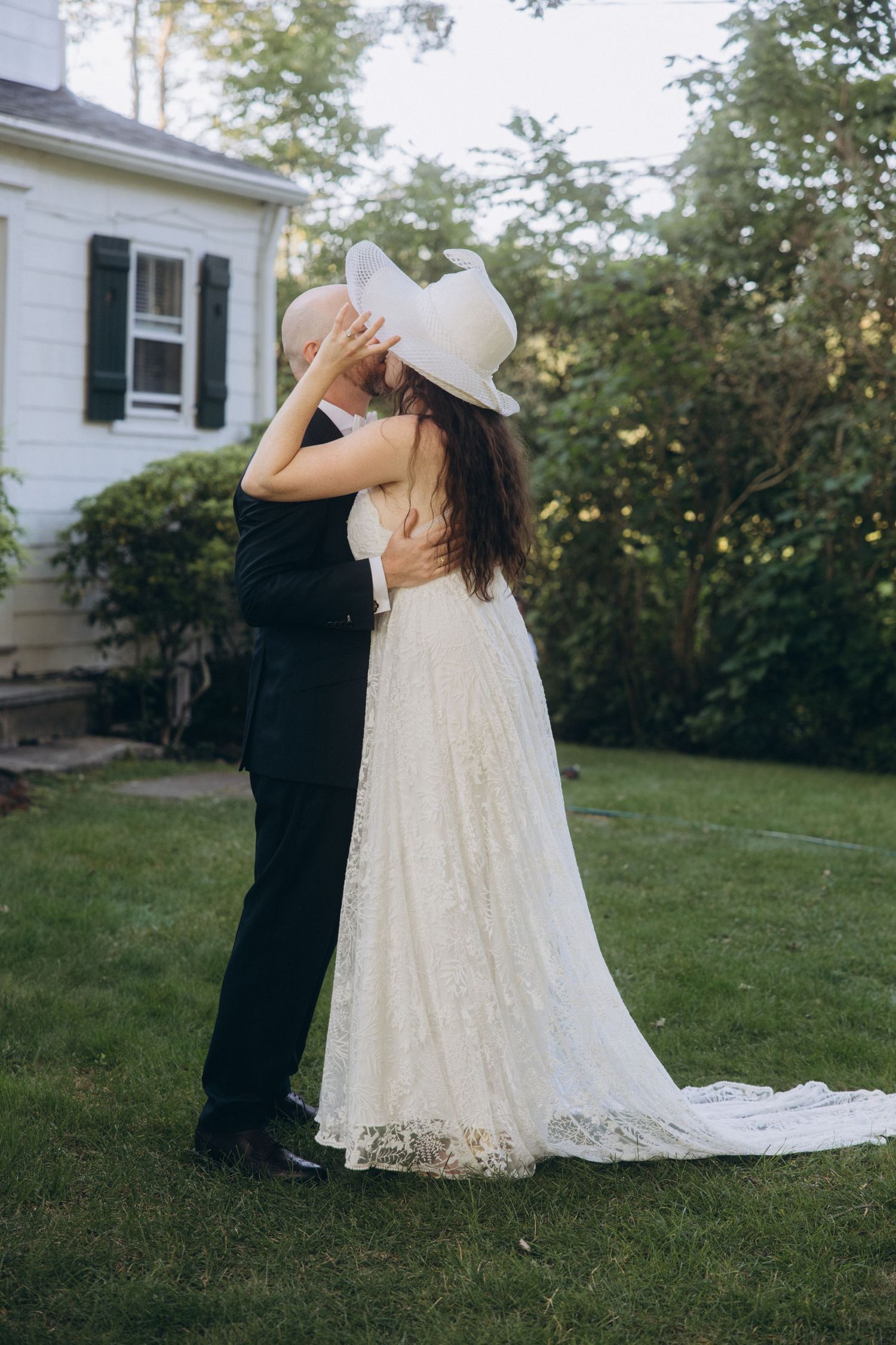 Romantic outdoor wedding portrait with couple embracing in front of white house with flowing lace dress.