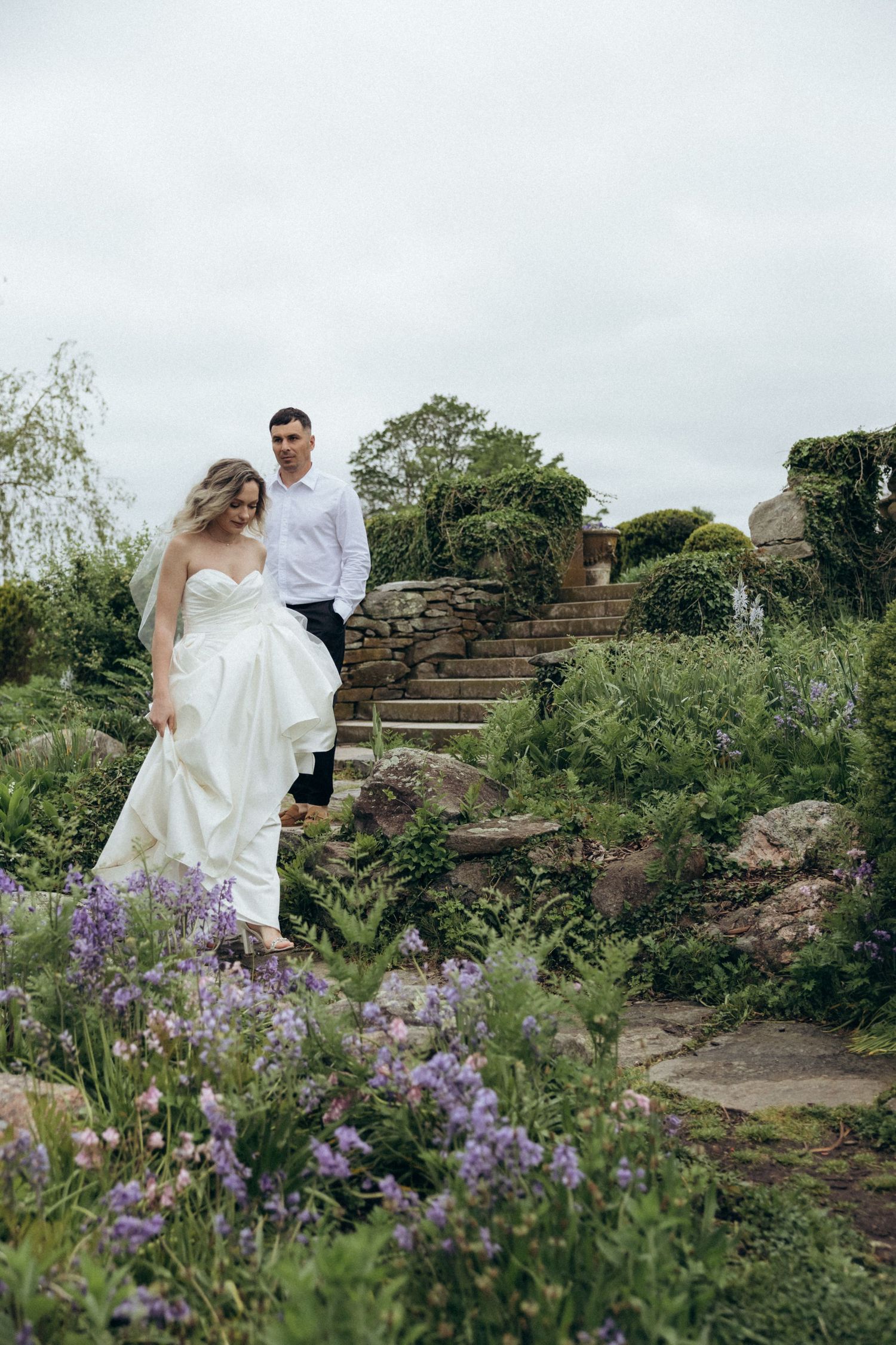 Wedding couple posing on stone steps surrounded by purple wildflowers.
